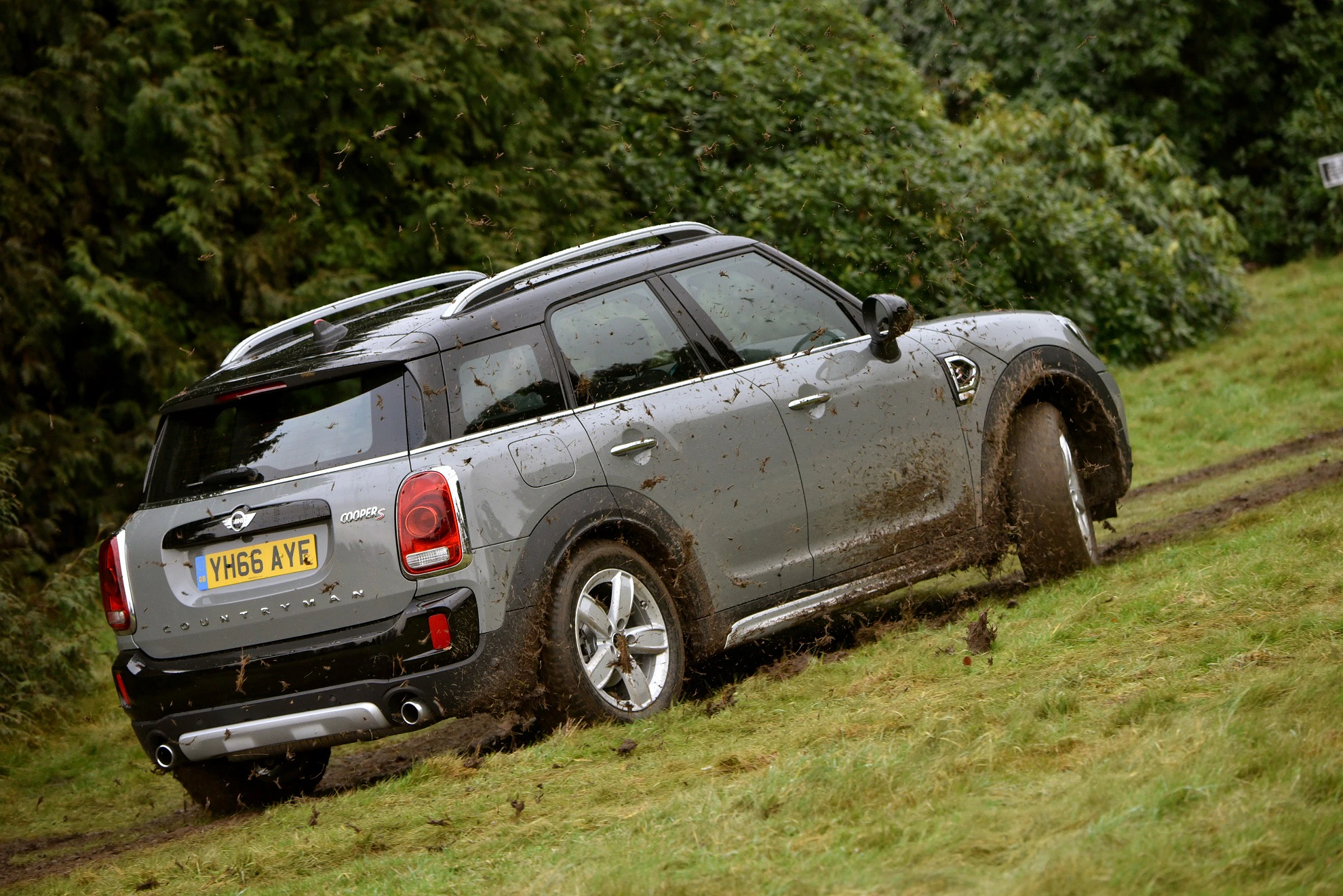 Ein grauer Mini-SUV fährt durch schlammiges Gelände, während Erde aufspritzt. Im Hintergrund sind Bäume sichtbar.