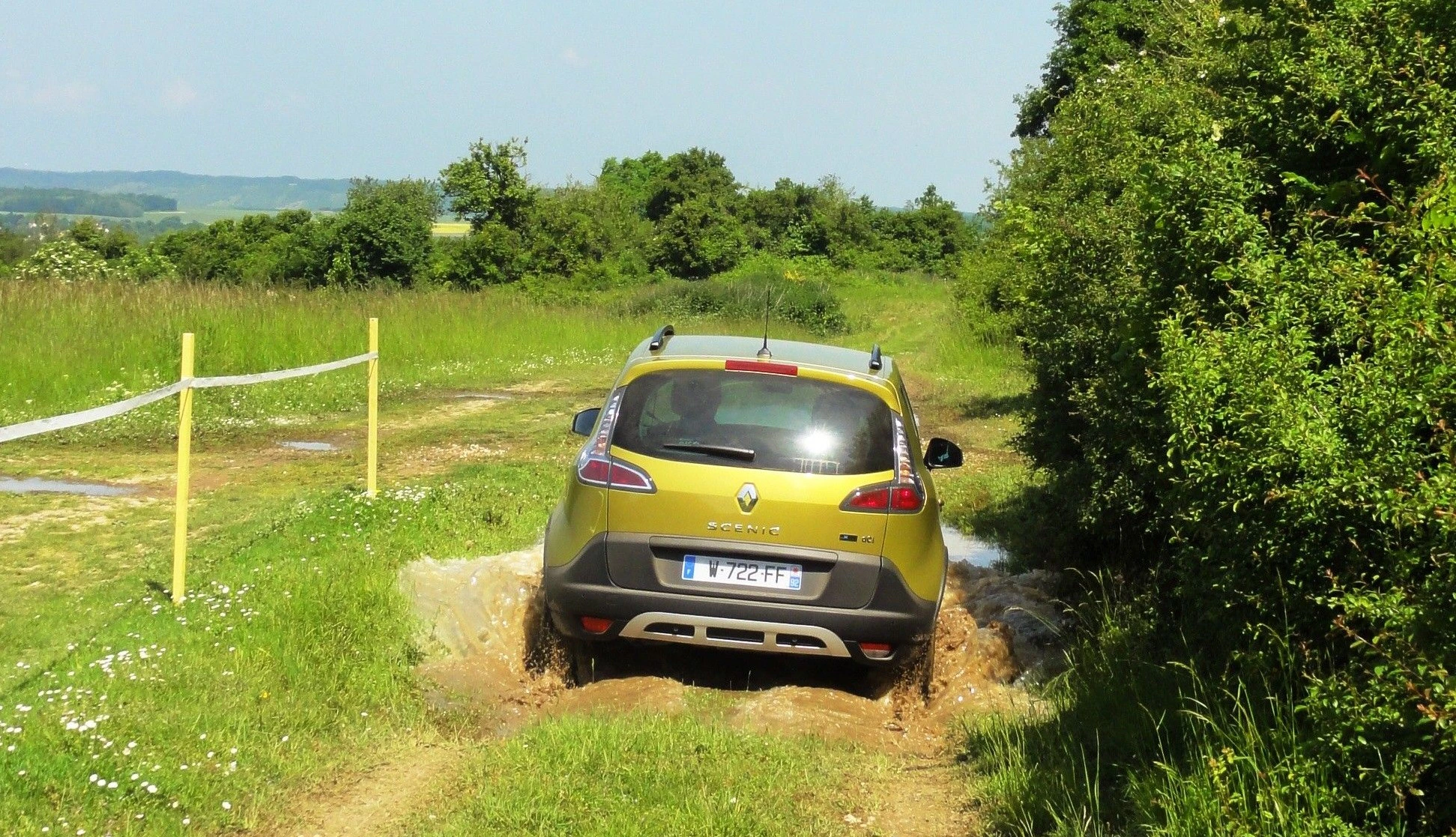 Gelb-grünes Auto fährt durch schlammige Pfütze auf einem ländlichen Weg, umgeben von grünem Grass und Büschen unter blauem Himmel.