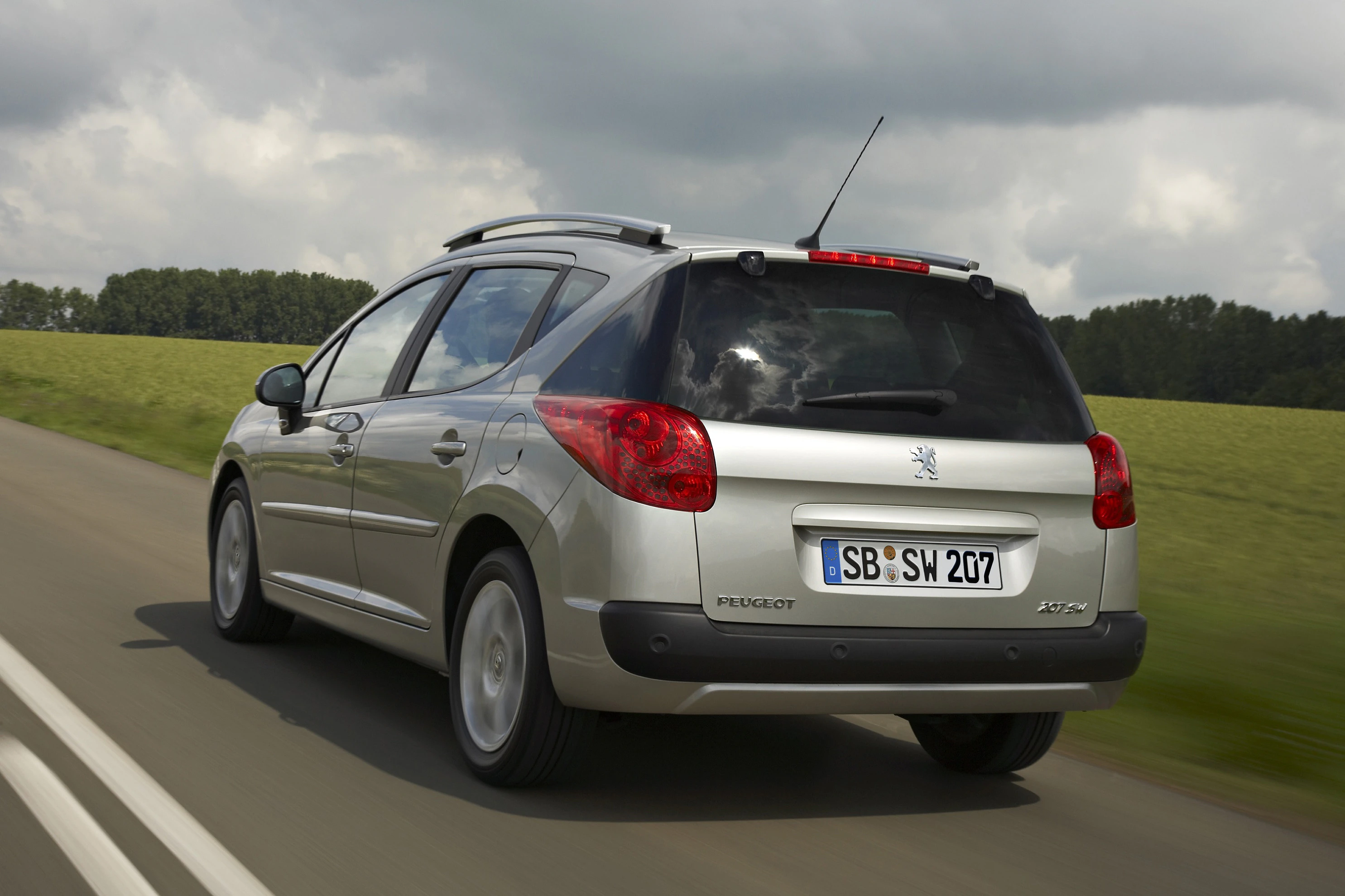 Silberner Peugeot 207 SW fährt auf einer Landstraße. Bewölkter Himmel und grüne Felder im Hintergrund.