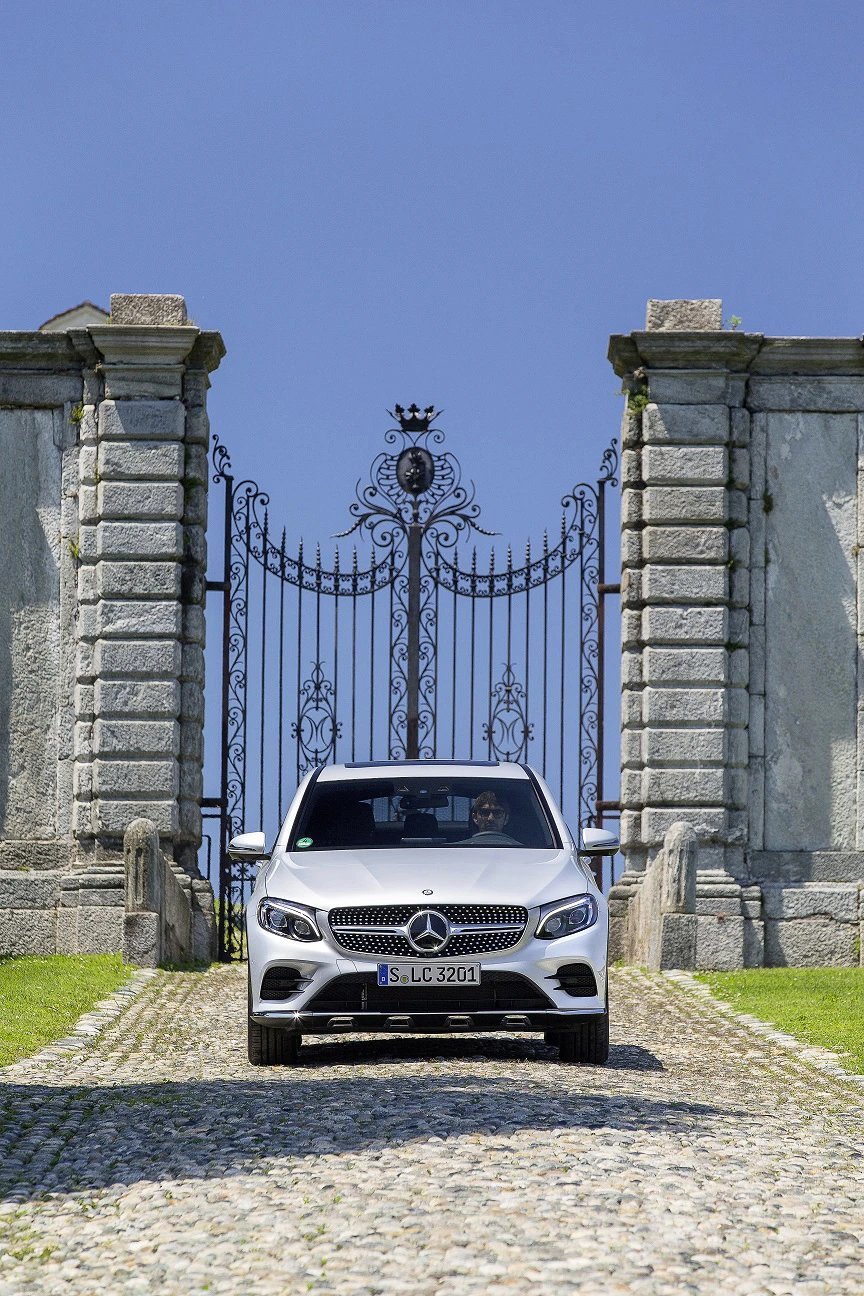 Silberner Mercedes fährt durch antikes Steintor mit verziertem Eisentor, auf gepflastertem Weg, grüner Rasen und blauer Himmel im Hintergrund.