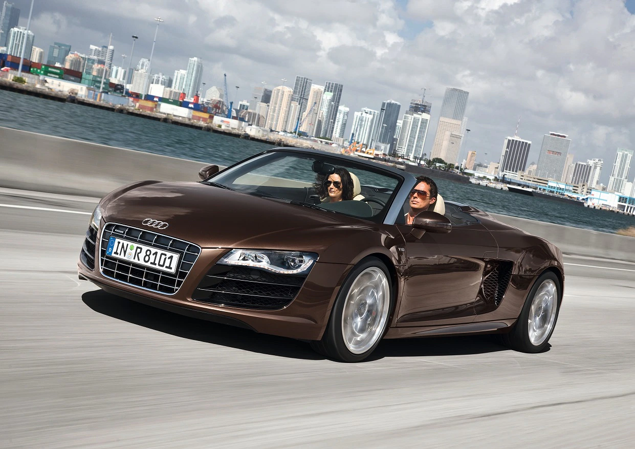 Zwei Personen fahren in einem braunen Audi R8 Spyder auf einer Küstenstraße, im Hintergrund eine Stadt mit Wolkenkratzern und Hafen.