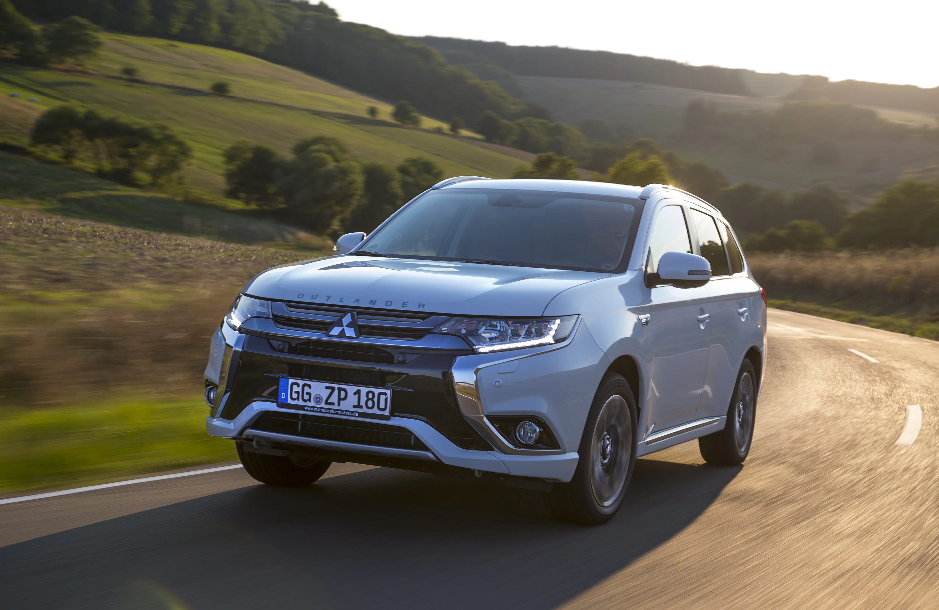 Ein silberner Mitsubishi Outlander fährt bei Sonnenuntergang auf einer Landstraße durch hügelige Landschaft.