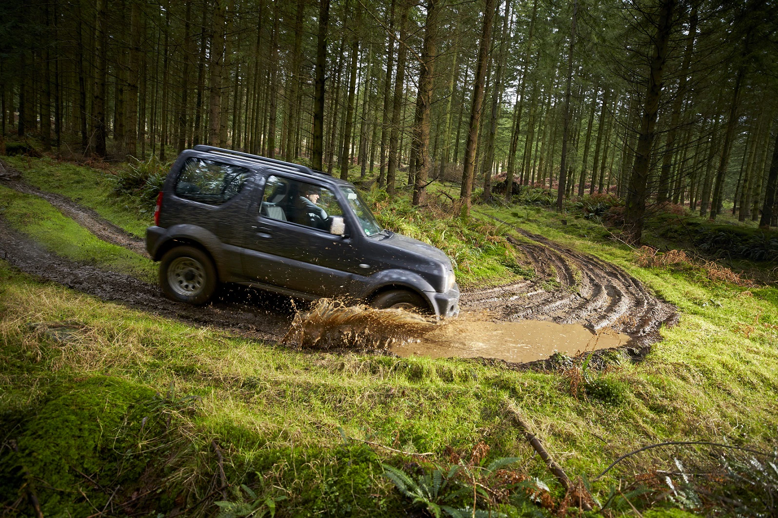 Geländewagen fährt durch schlammige Spur im Wald, Spritzwasser, umgeben von hohen Bäumen und grünem Moos.