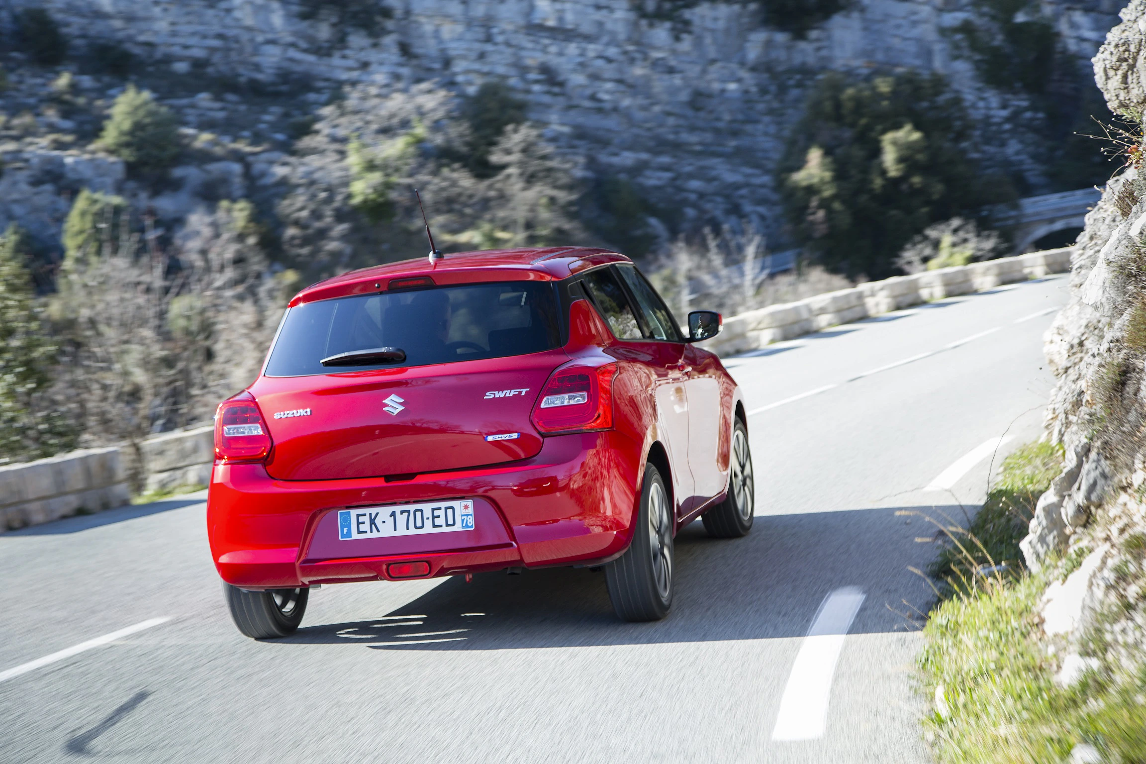 Roter Suzuki Swift fährt auf kurviger Bergstraße bei sonnigem Wetter, umgeben von Felsen und Bäumen.