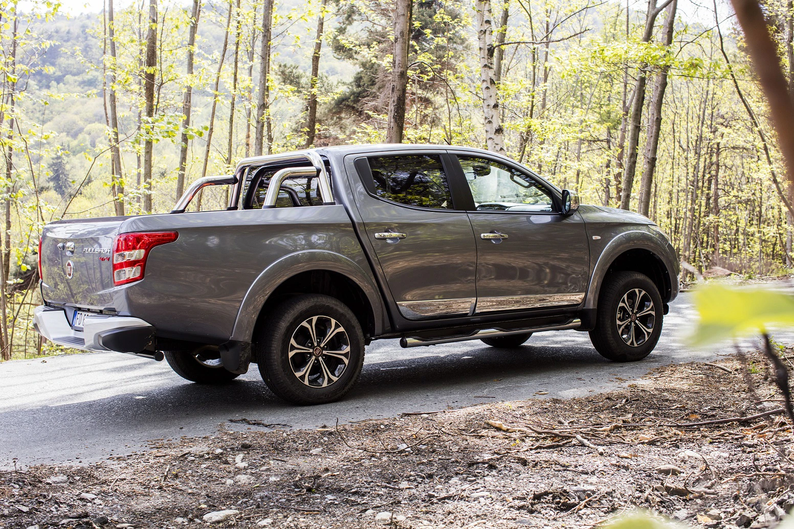 Grau-silberner Pickup-Truck fährt auf einer Waldstraße, umgeben von Bäumen und natürlicher Landschaft.