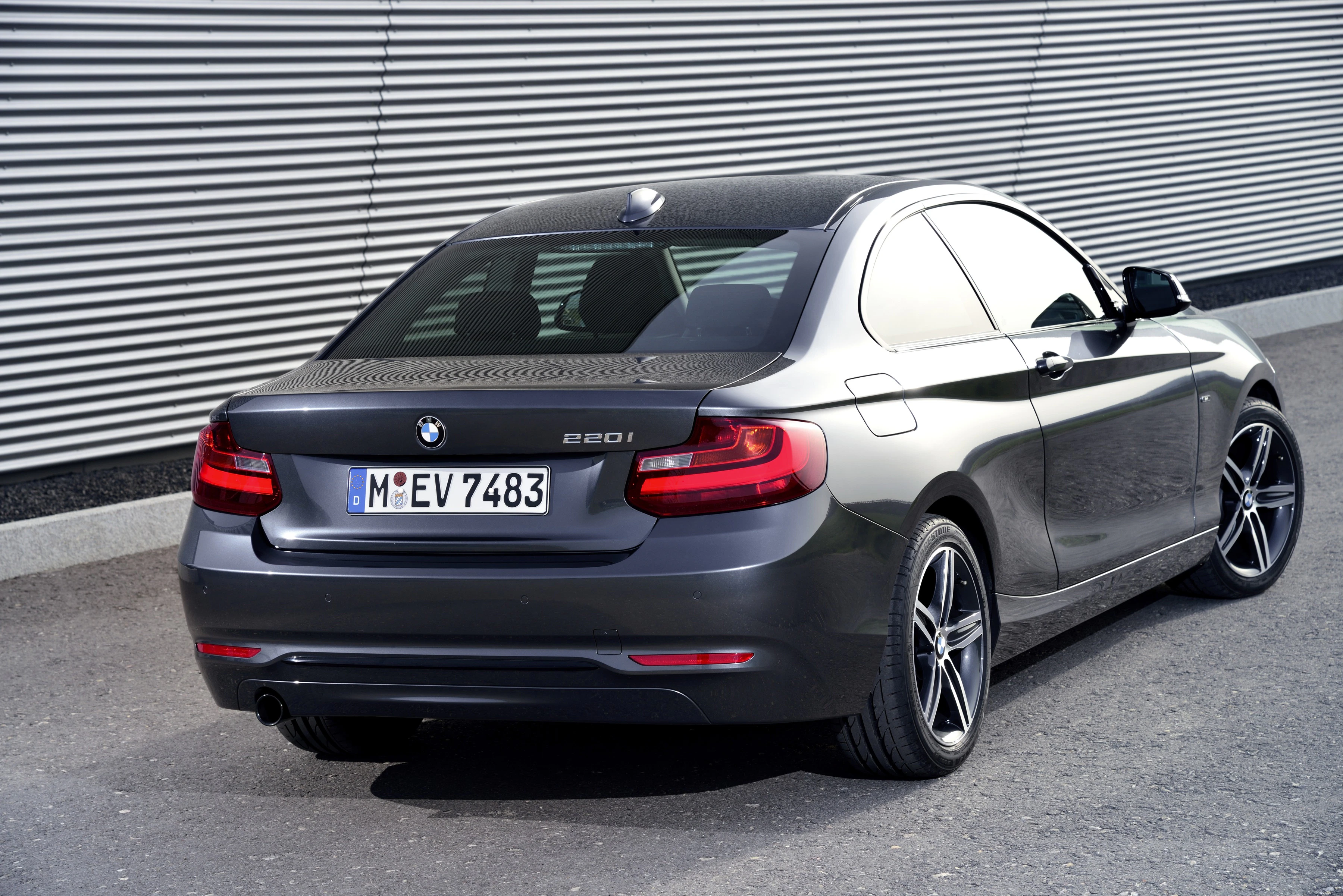 Dunkelgrauer BMW 220i auf Asphalt vor einer grauen, geriffelten Wand, von hinten fotografiert.
