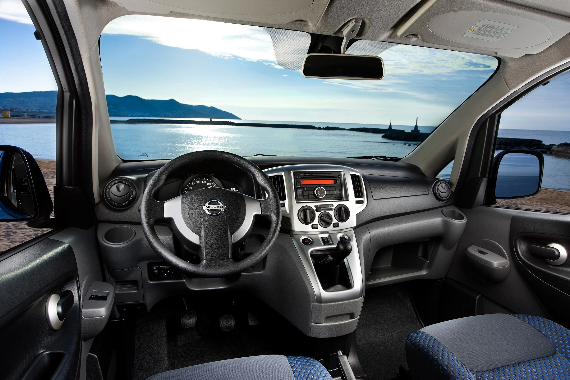 Cockpit eines Nissan-Fahrzeugs mit Blick aufs Meer. Moderne Armaturen und Lenkrad, dahinter ein sonniger Horizont mit Bergen und Wasser.
