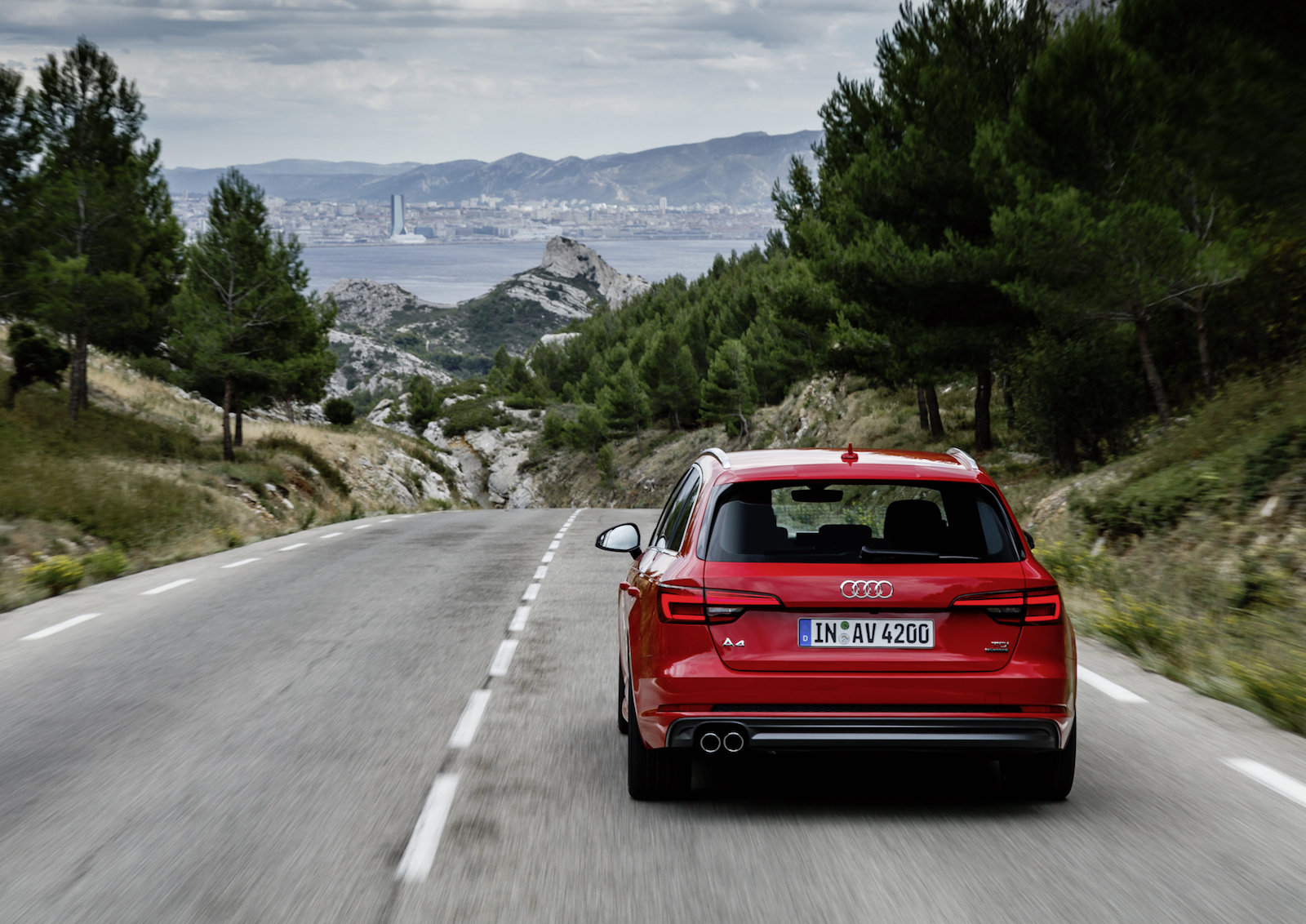 Roter Audi auf einer Landstraße, umgeben von Bäumen, mit Blick auf eine Küstenstadt und Berge im Hintergrund.