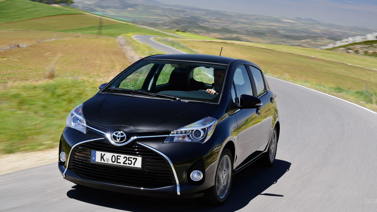Schwarzer Toyota Yaris fährt auf einer kurvenreichen Landstraße durch eine malerische Hügellandschaft bei sonnigem Wetter.
