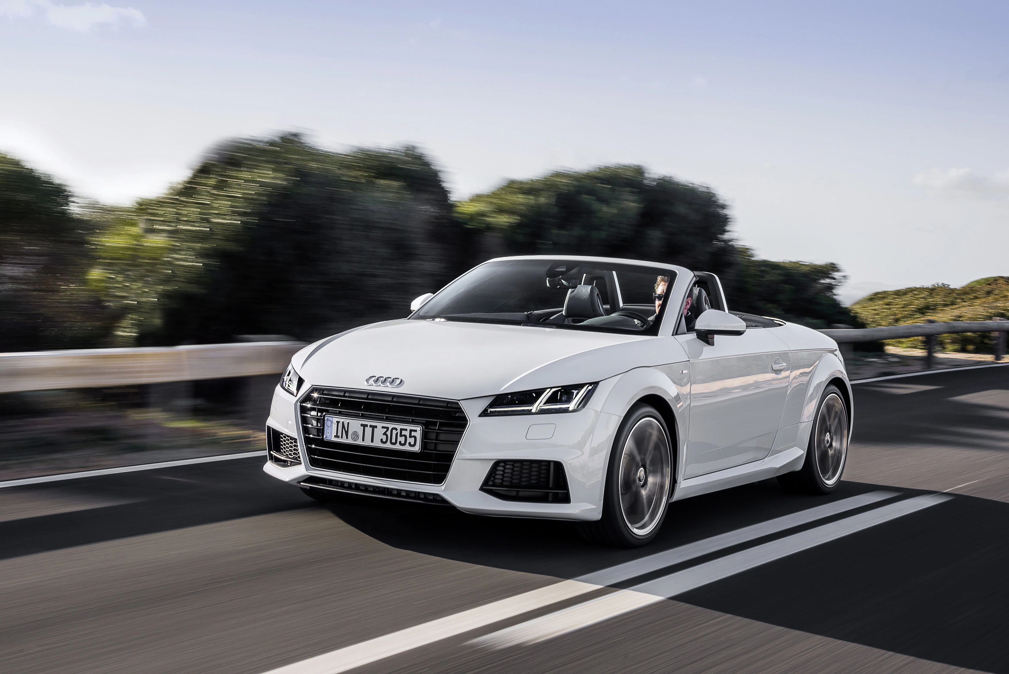 Weißer Audi TT Cabrio fährt auf einer Landstraße, umgeben von Bäumen. Es ist ein sonniger Tag mit klarem Himmel.