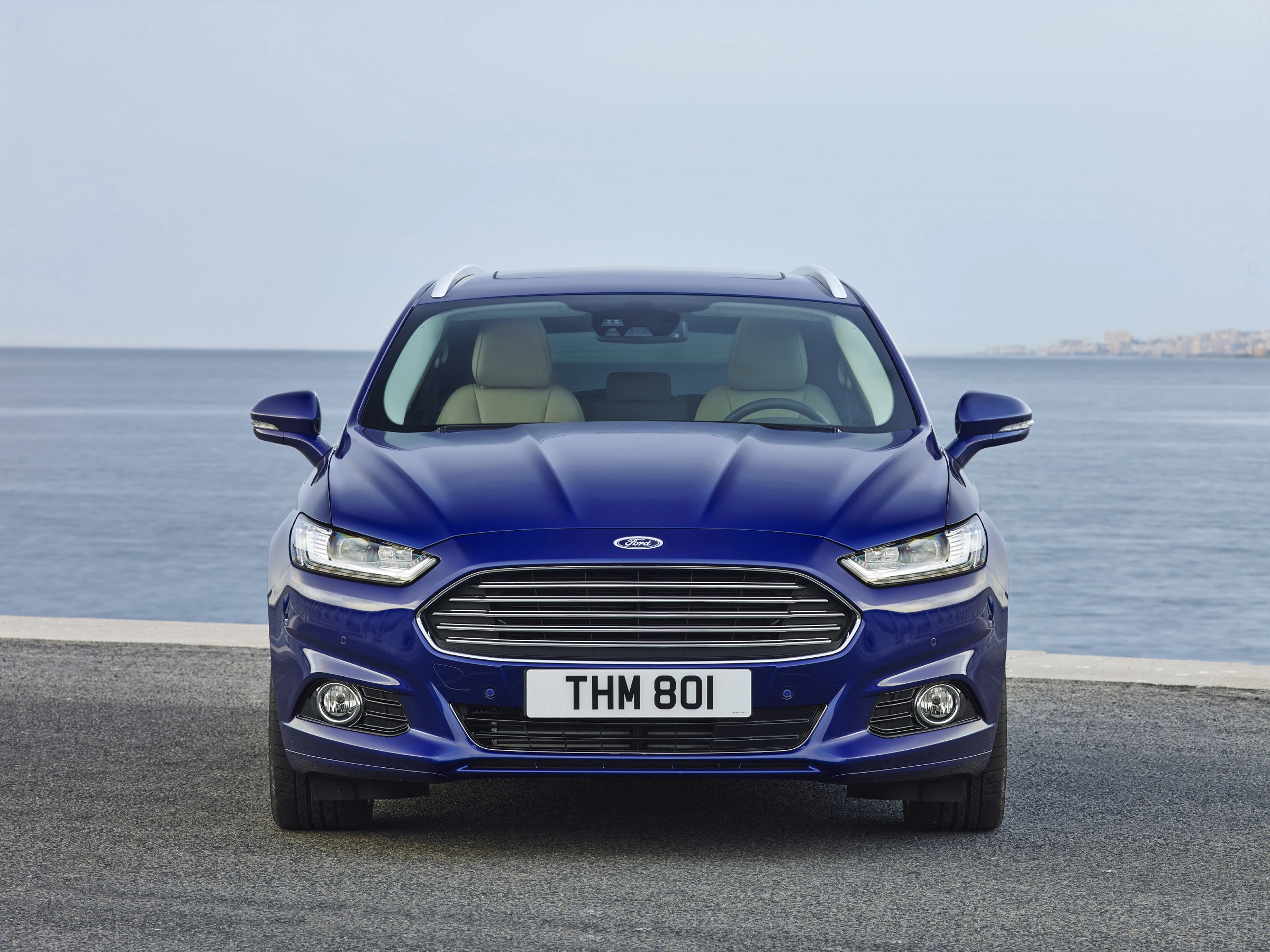 Blaues Auto der Marke Ford steht auf einer Küstenstraße vor einem ruhigen Meer, mit klarem Himmel im Hintergrund.
