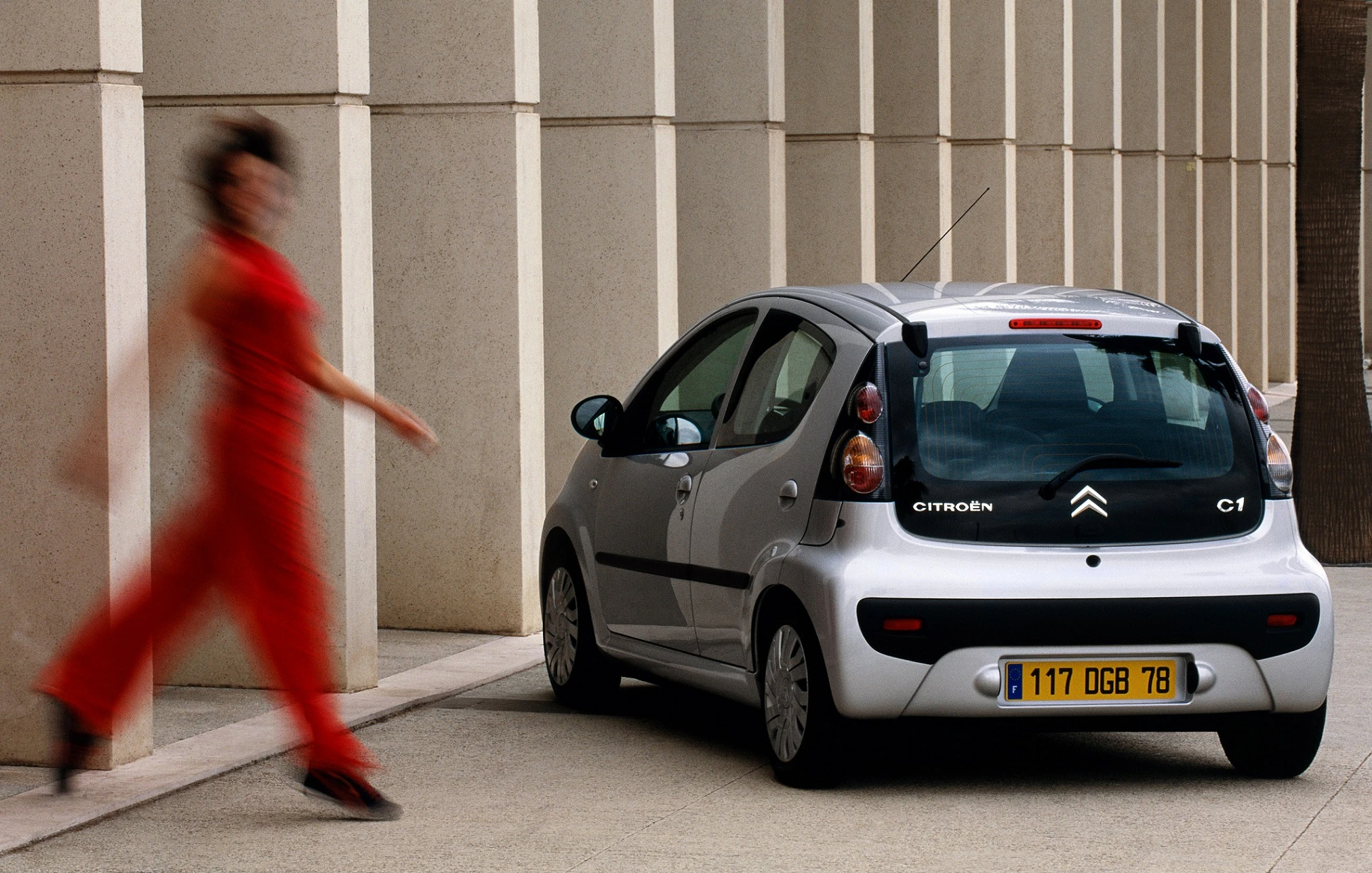 Silberner Citroën C1 geparkt neben einer modernen Gebäudewand; eine Person in rotem Outfit geht schnell im Vordergrund vorbei.