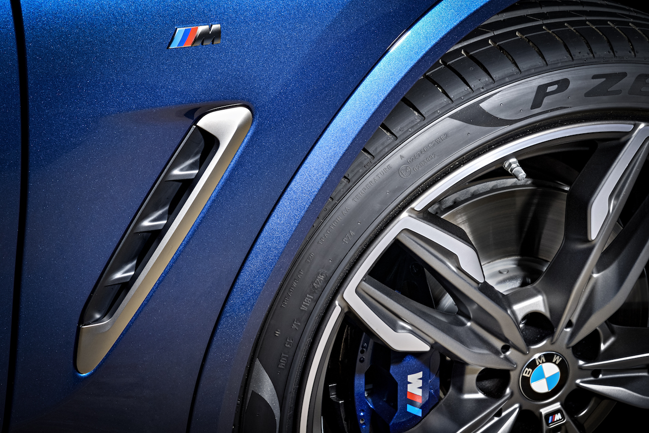 Nahaufnahme eines blauen BMW-Fahrzeugs mit M-Logo, zeigt Design-Details und Reifen mit Felge, unterstreicht sportliches Aussehen.