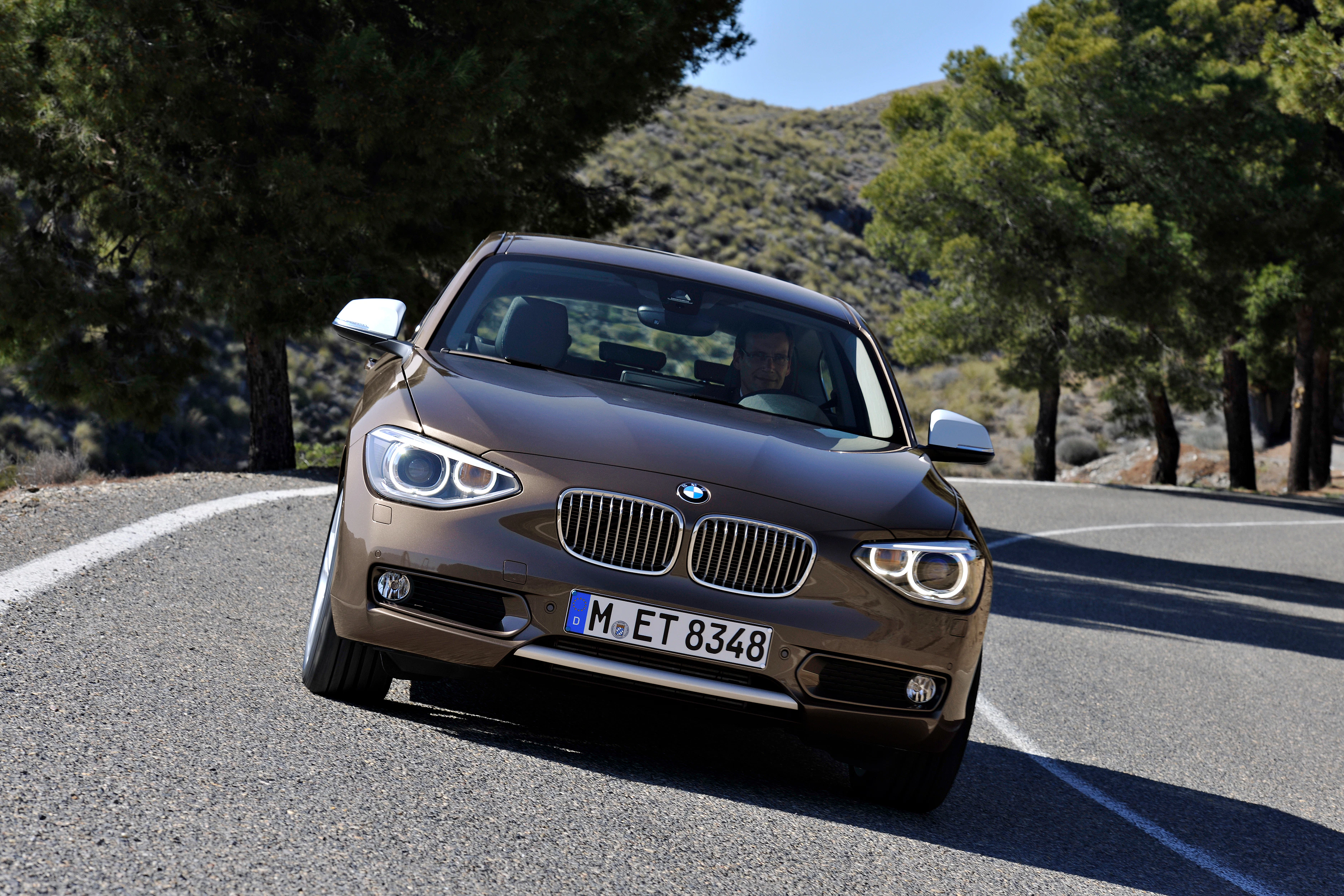 Ein brauner BMW fährt auf einer kurvigen Landstraße, umgeben von Bäumen und Hügeln.