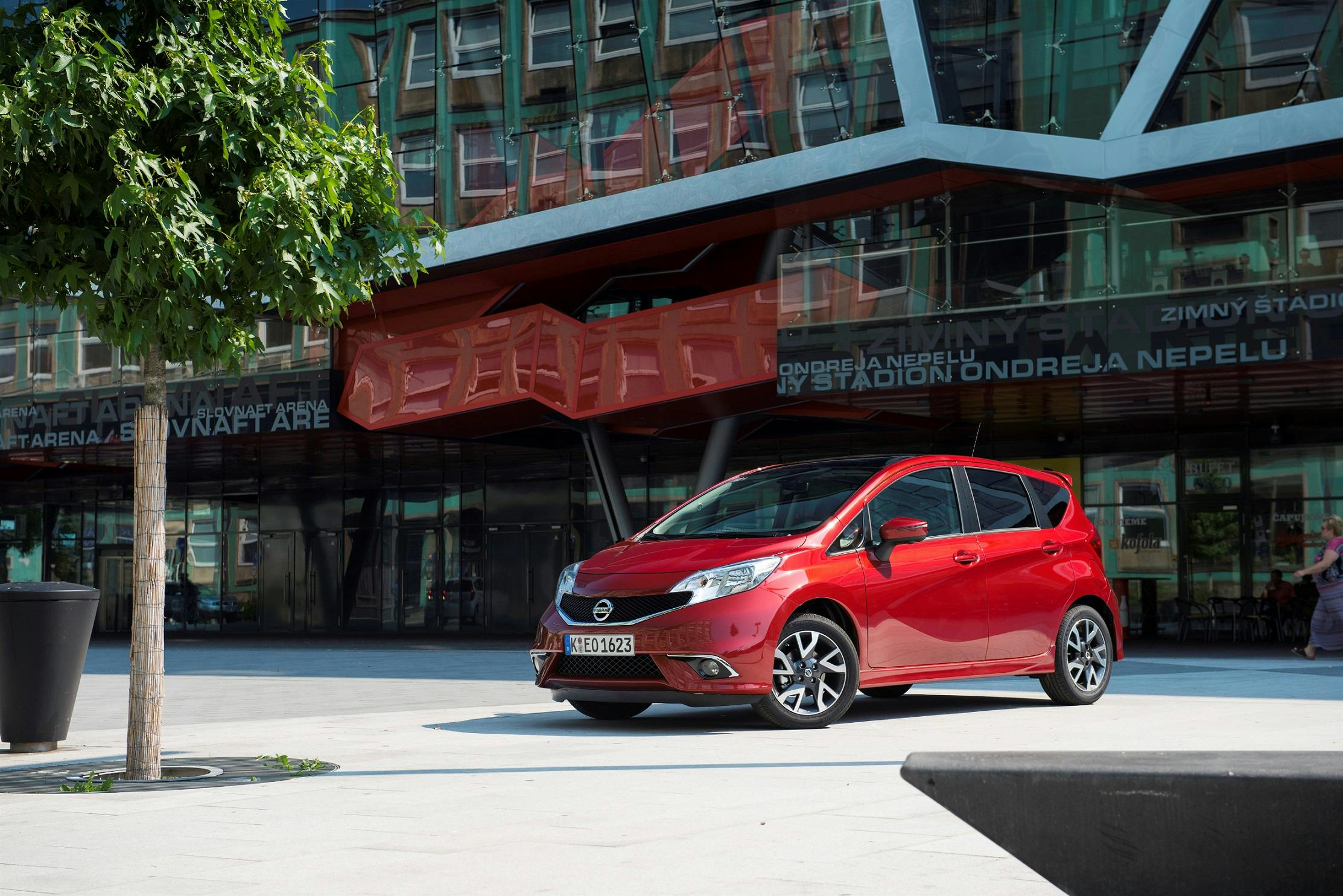 Rotes Auto parkt vor einem modernen Gebäude mit Glasfassade; im Vordergrund ein Baum, im Hintergrund reflektierende Fenster.