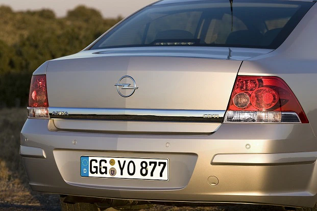 Rückansicht eines beigen Opel Vectra mit deutschem Kennzeichen, rotem Rücklicht und grünlicher Naturkulisse im Hintergrund.