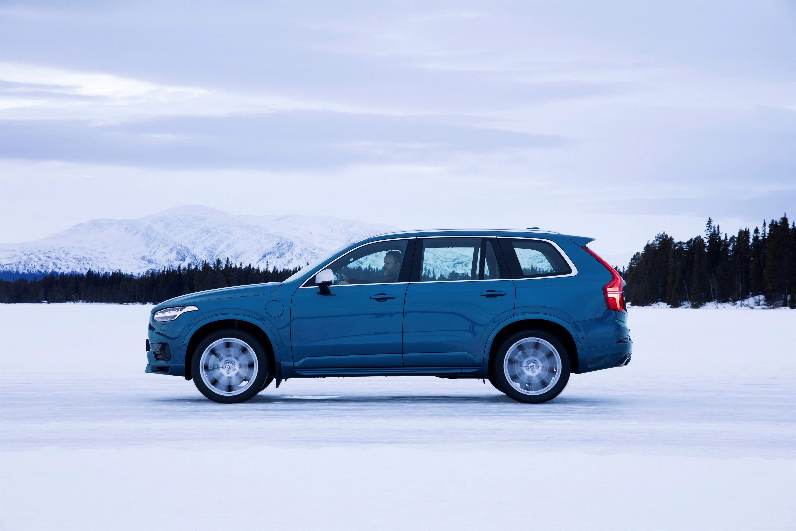 Blauer SUV steht auf einer verschneiten, weiten Fläche mit Wald und Bergen im Hintergrund unter einem bewölkten Himmel.