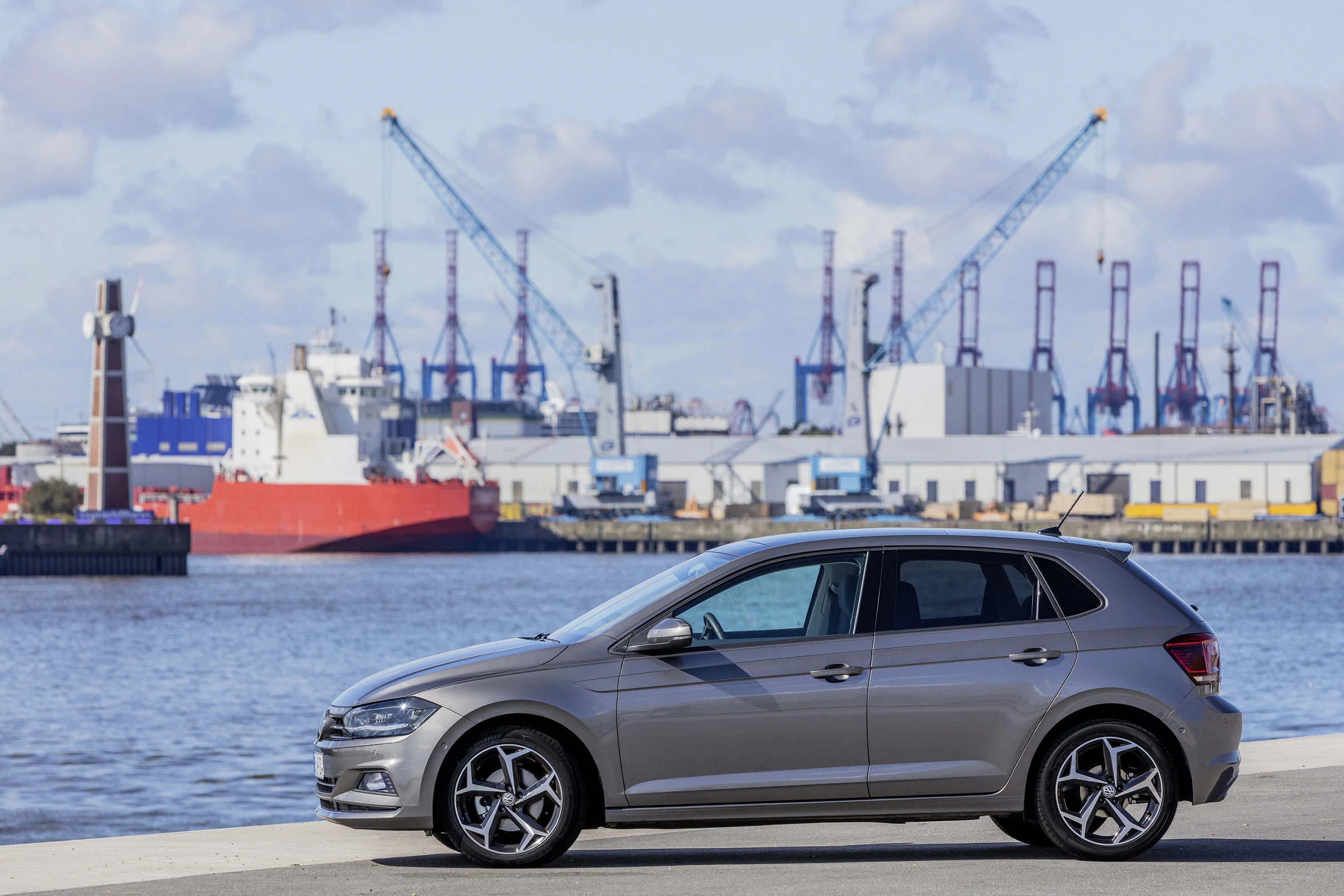 Auto vor einem Hafen mit Kränen und Schiffen im Hintergrund. Blauer Himmel und Wolken ergänzen die Szenerie.