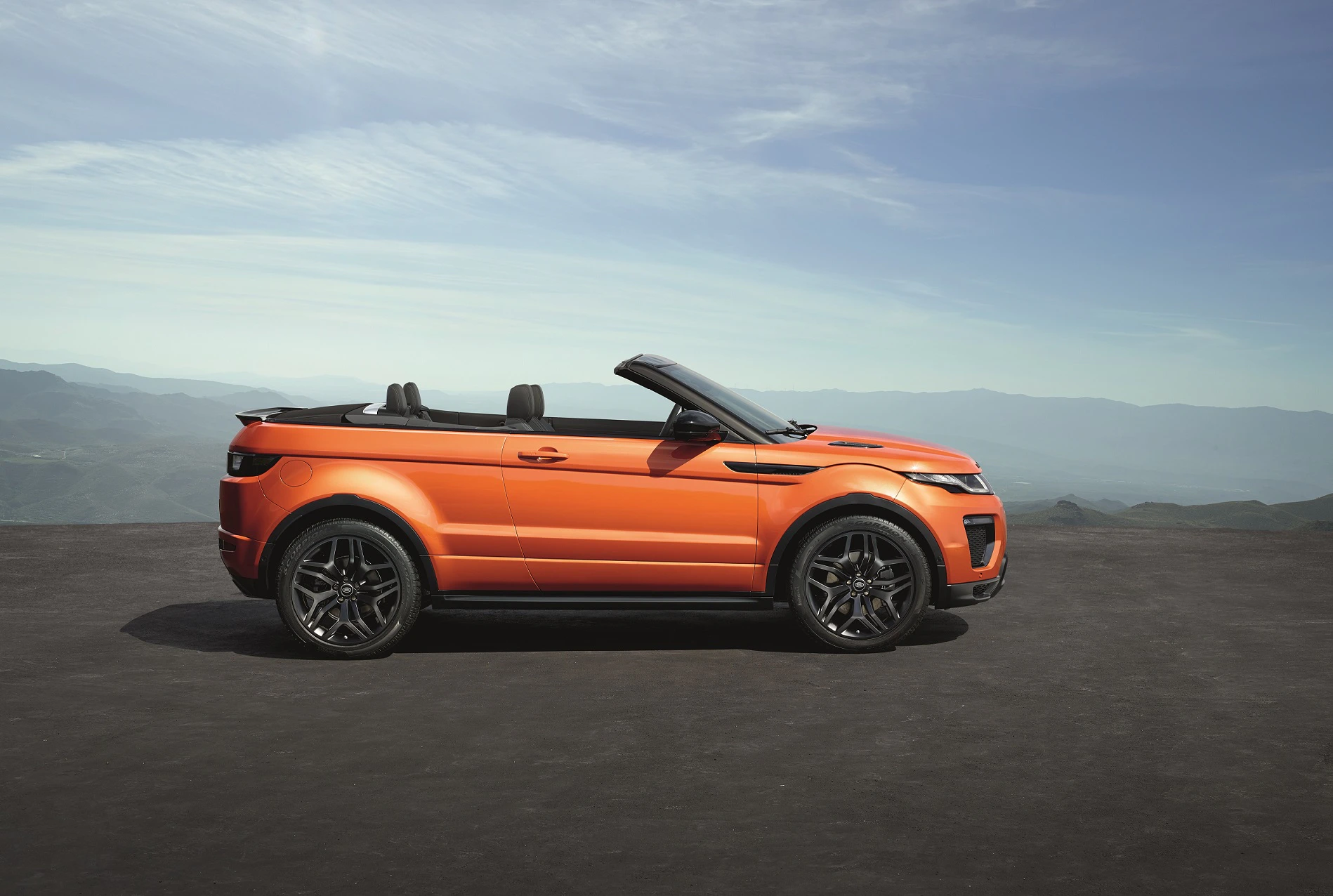 Orangefarbenes Cabrio-Geländewagen mit offenem Verdeck, geparkt auf einer Straße. Im Hintergrund sind Berge und Himmel zu sehen.