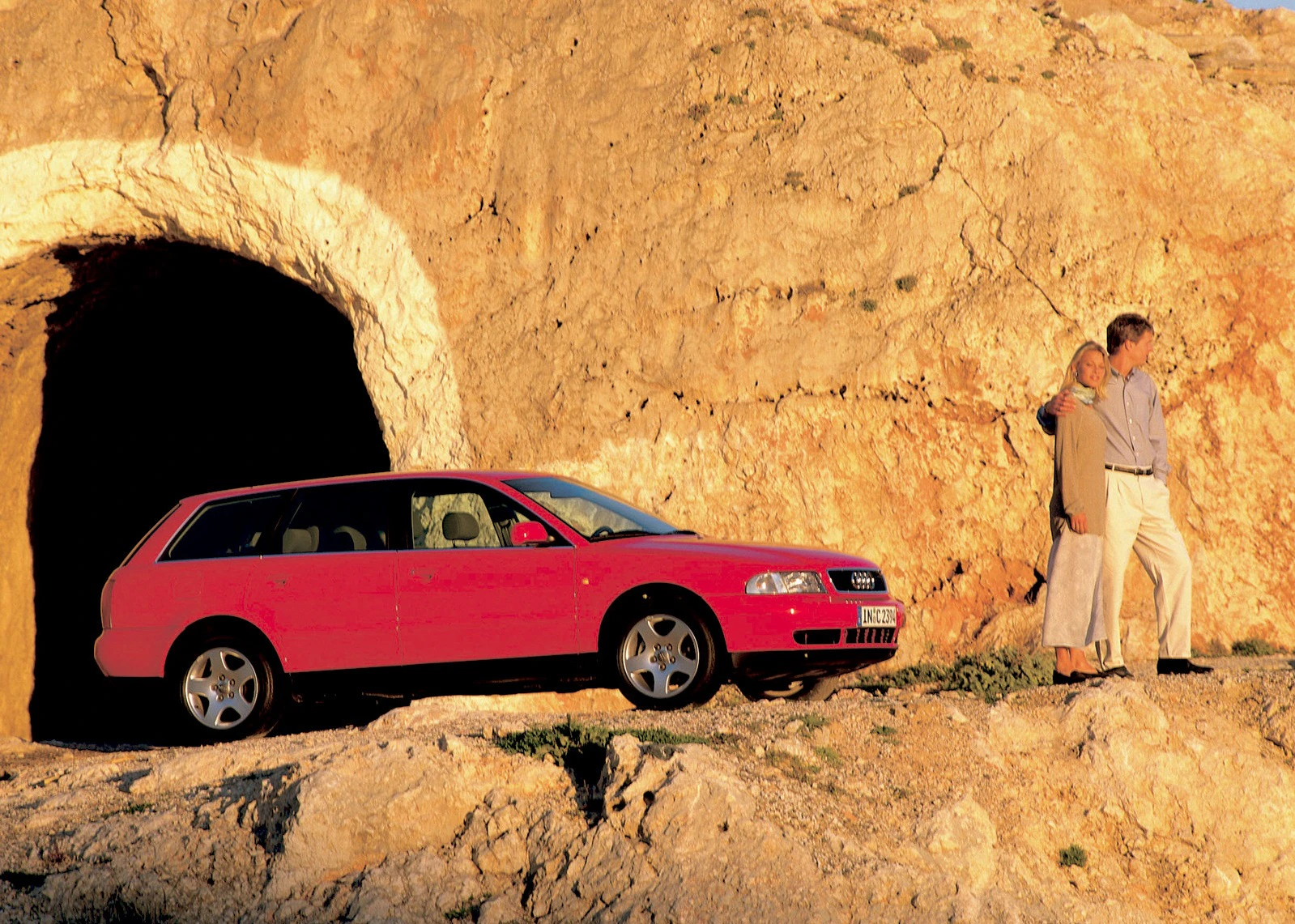 Roter Audi Kombi vor einer Felswand mit Höhle; daneben stehen zwei Personen, die in die Ferne schauen.