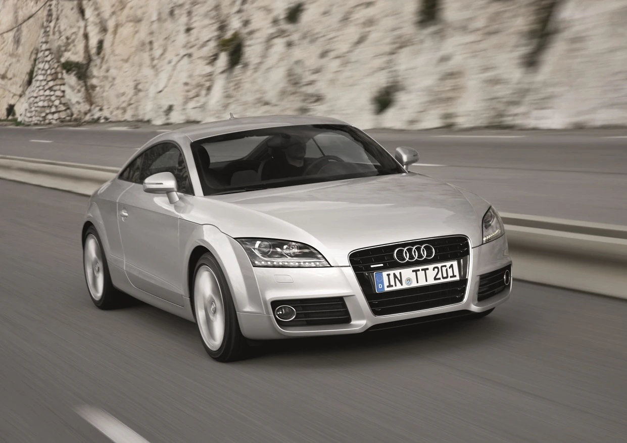 Silberner Audi TT fährt auf einer kurvigen Straße entlang einer Felswand. Sportliches Design, dynamische Kurven.