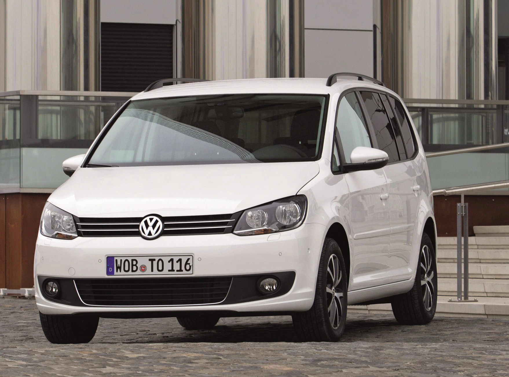 Weißer Volkswagen Touran steht auf einer gepflasterten Fläche vor einem modernen Gebäude.