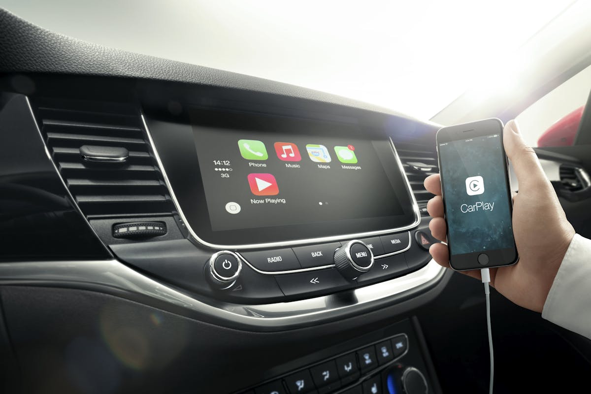 Innenraum eines Autos mit aktiviertem Apple CarPlay. Eine Hand hält ein Smartphone, das über ein Kabel mit dem Infotainment-Bildschirm verbunden ist.
