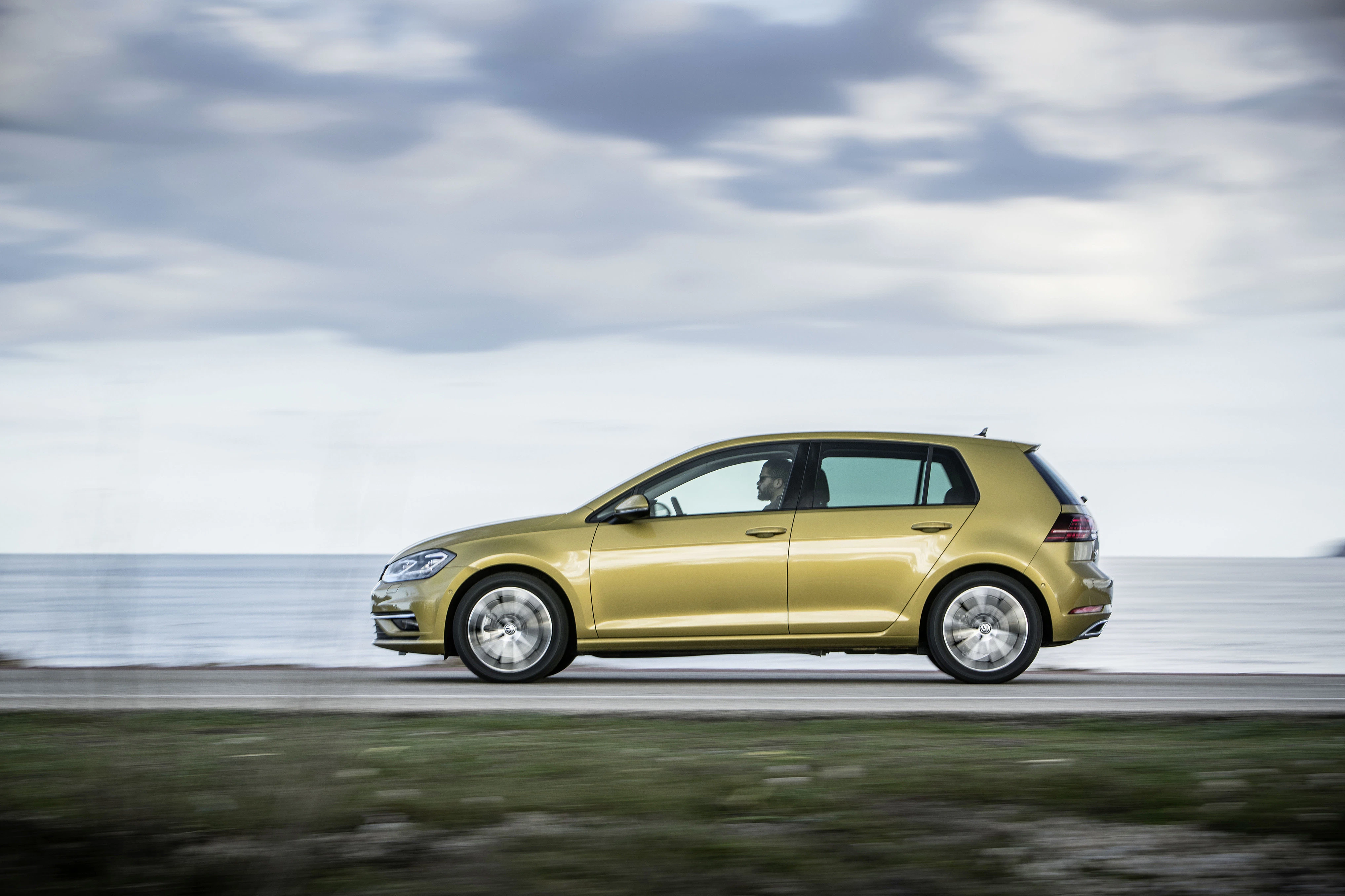 Goldener VW Golf fährt bei bewölktem Himmel am Meer entlang.