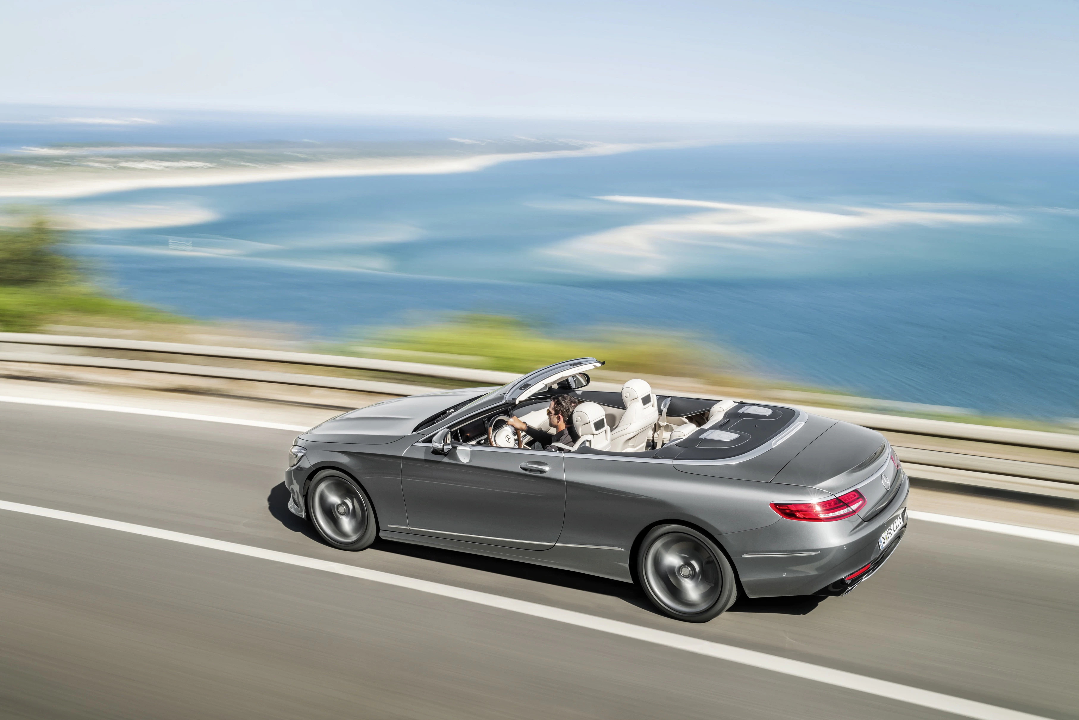 Graues Cabrio fährt auf einer Küstenstraße. Im Hintergrund sind das blaue Meer und der Himmel zu sehen. Personen sitzen im Auto.