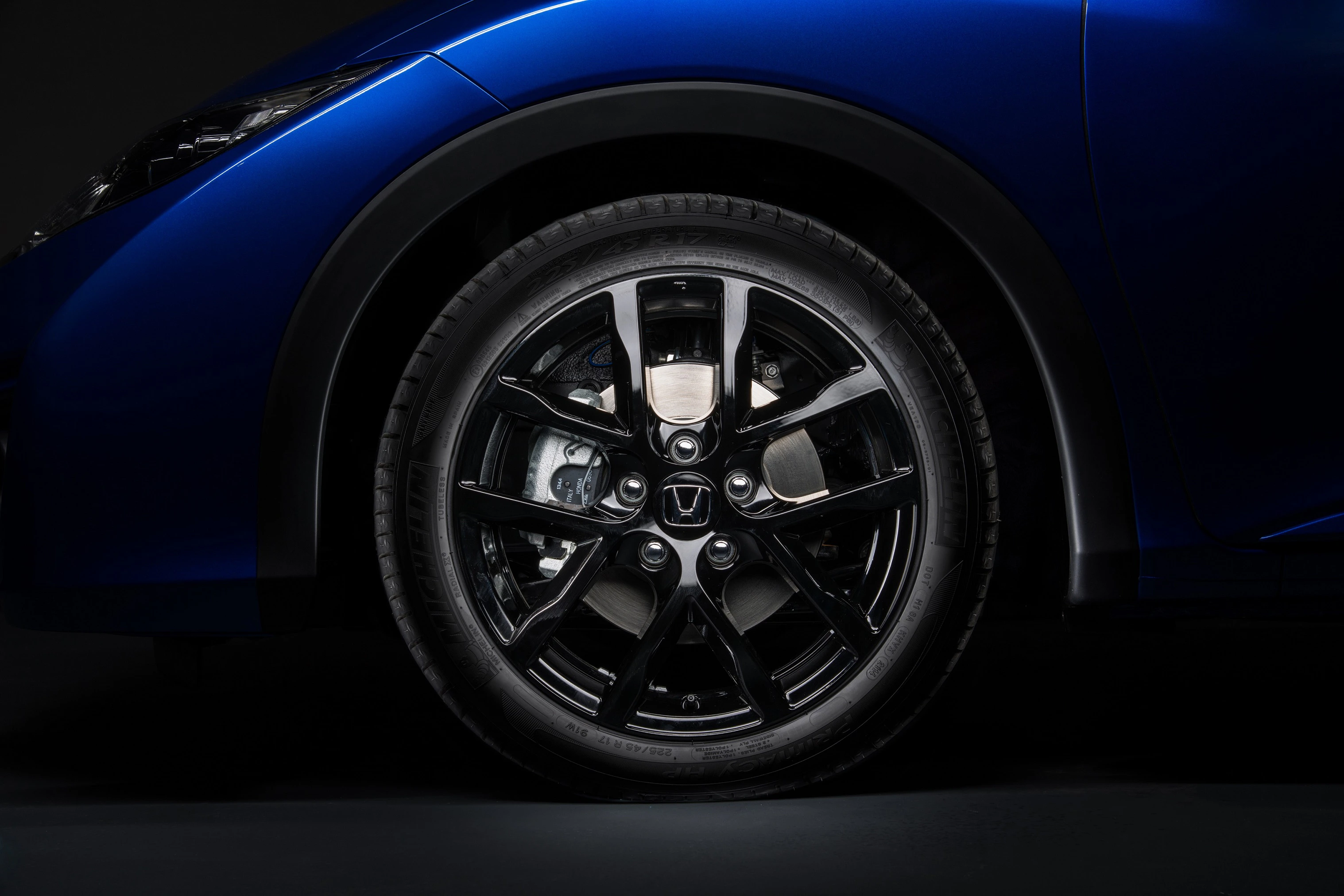 Nahaufnahme eines glänzenden schwarzen Autoreifens mit silbernen Felgen an einem blauen Fahrzeug, vor dunklem Hintergrund.