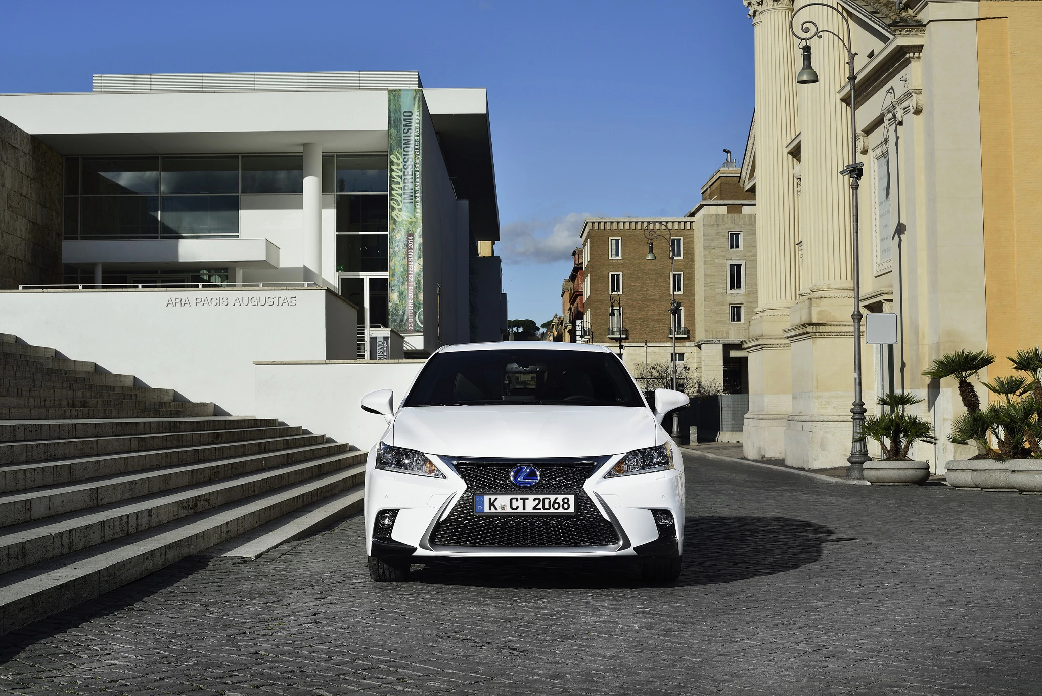 Weißer Lexus steht auf gepflasterter Straße vor modernen und historischen Gebäuden mit Treppe und Laterne.