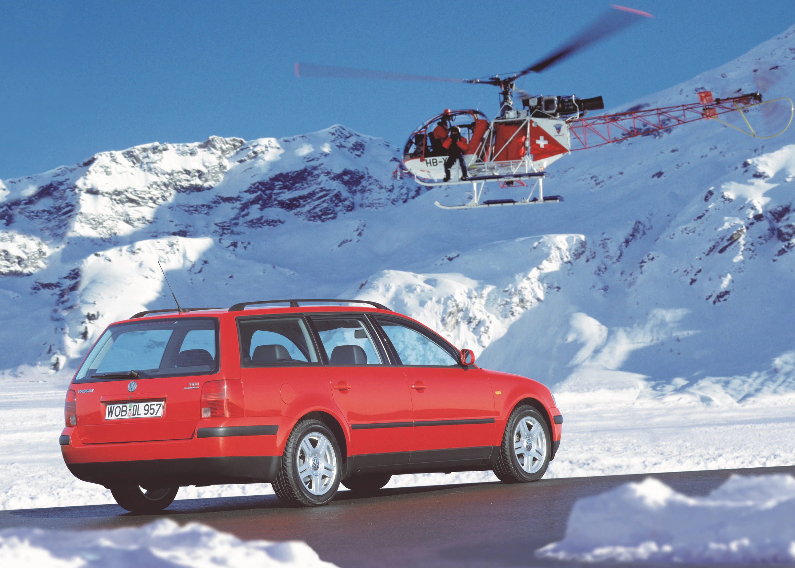 Roter Kombi auf verschneiter Bergstraße, Helikopter fliegt darüber, schneebedeckte Berge im Hintergrund.