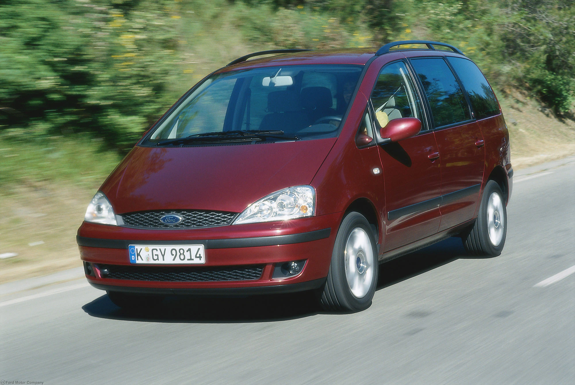 Roter Ford Galaxy fährt auf einer Landstraße, umgeben von Bäumen. Der Wagen hat ein deutsches Kennzeichen.