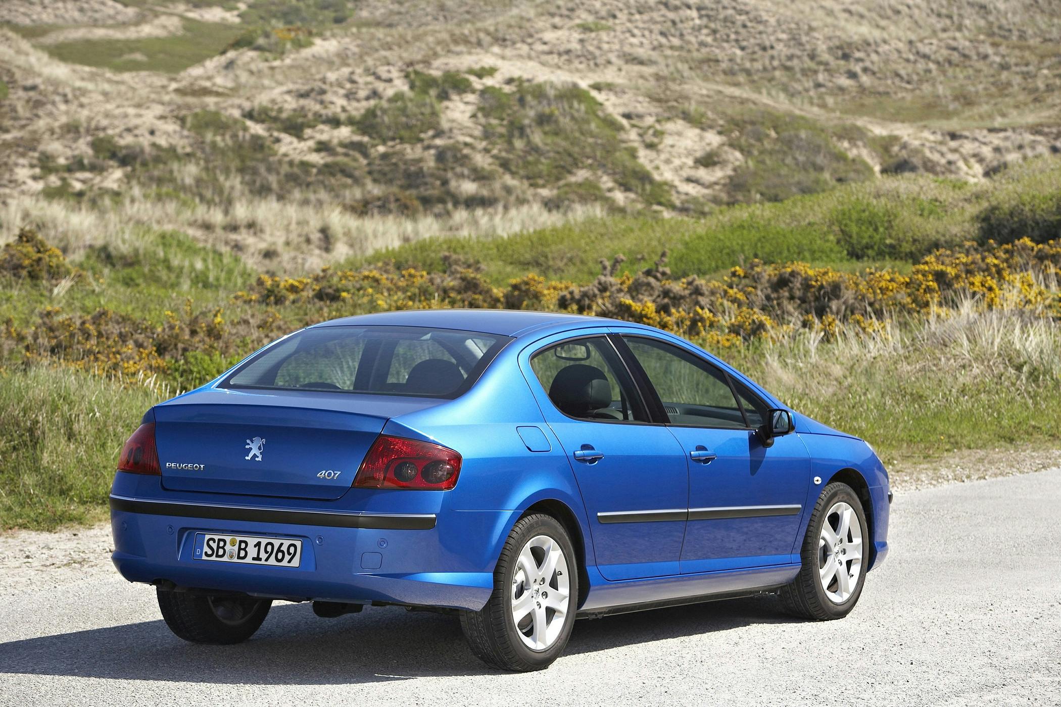 Blauer Peugeot 407 parkt auf einer ländlichen Straße, umgeben von sandigen Hügeln und spärlicher Vegetation. Rückansicht des Autos.