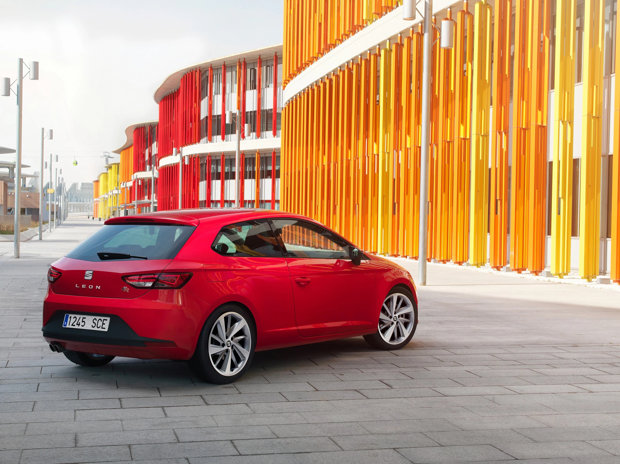 Roter Seat Leon parkt auf grauem Pflastersteinen vor moderner Architektur mit leuchtend orange-roten Fassaden.