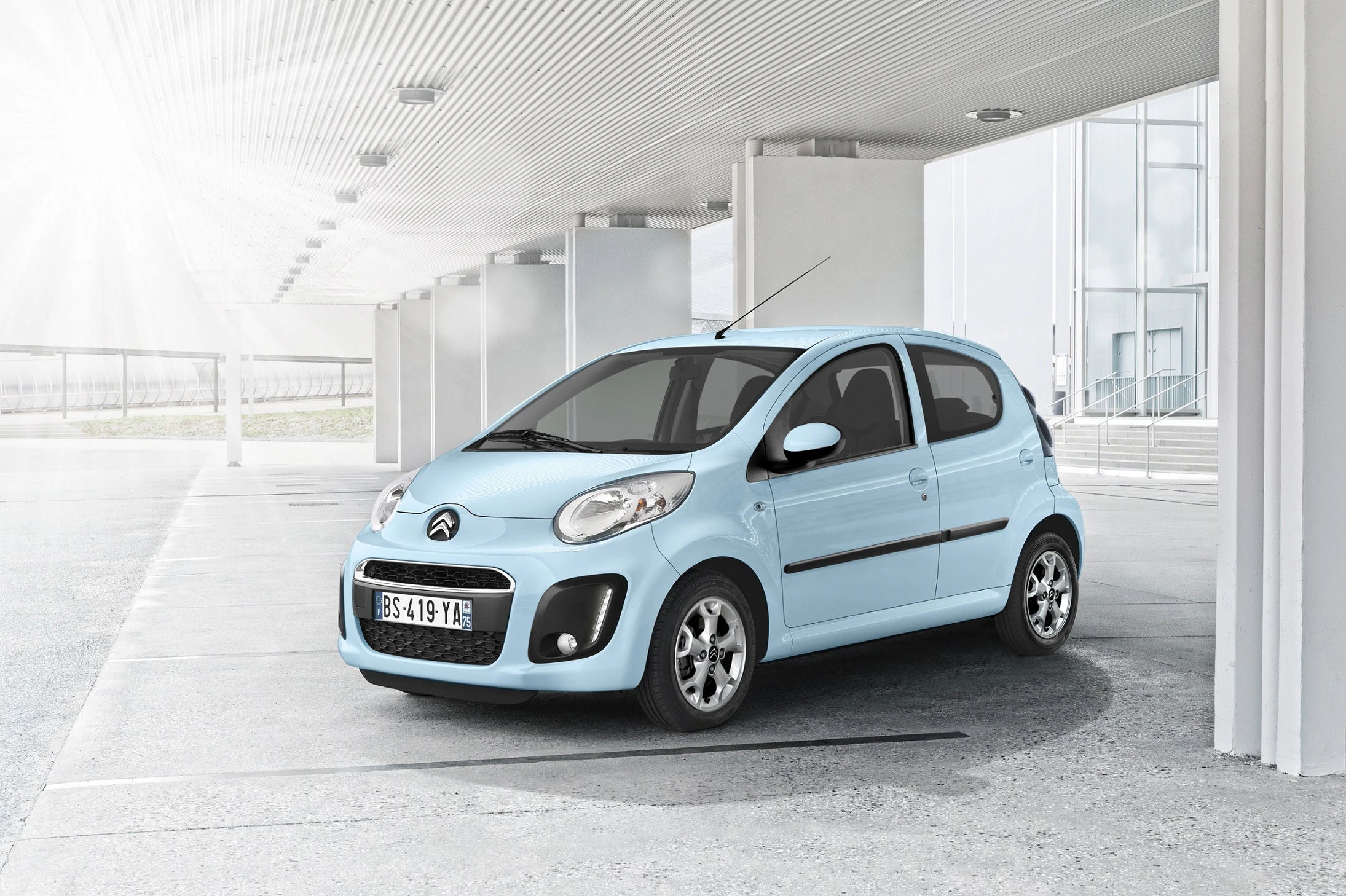 Heller blauer Citroën C1 steht unter einem modernen, überdachten Bereich mit Glasfenstern im Hintergrund bei Tageslicht.