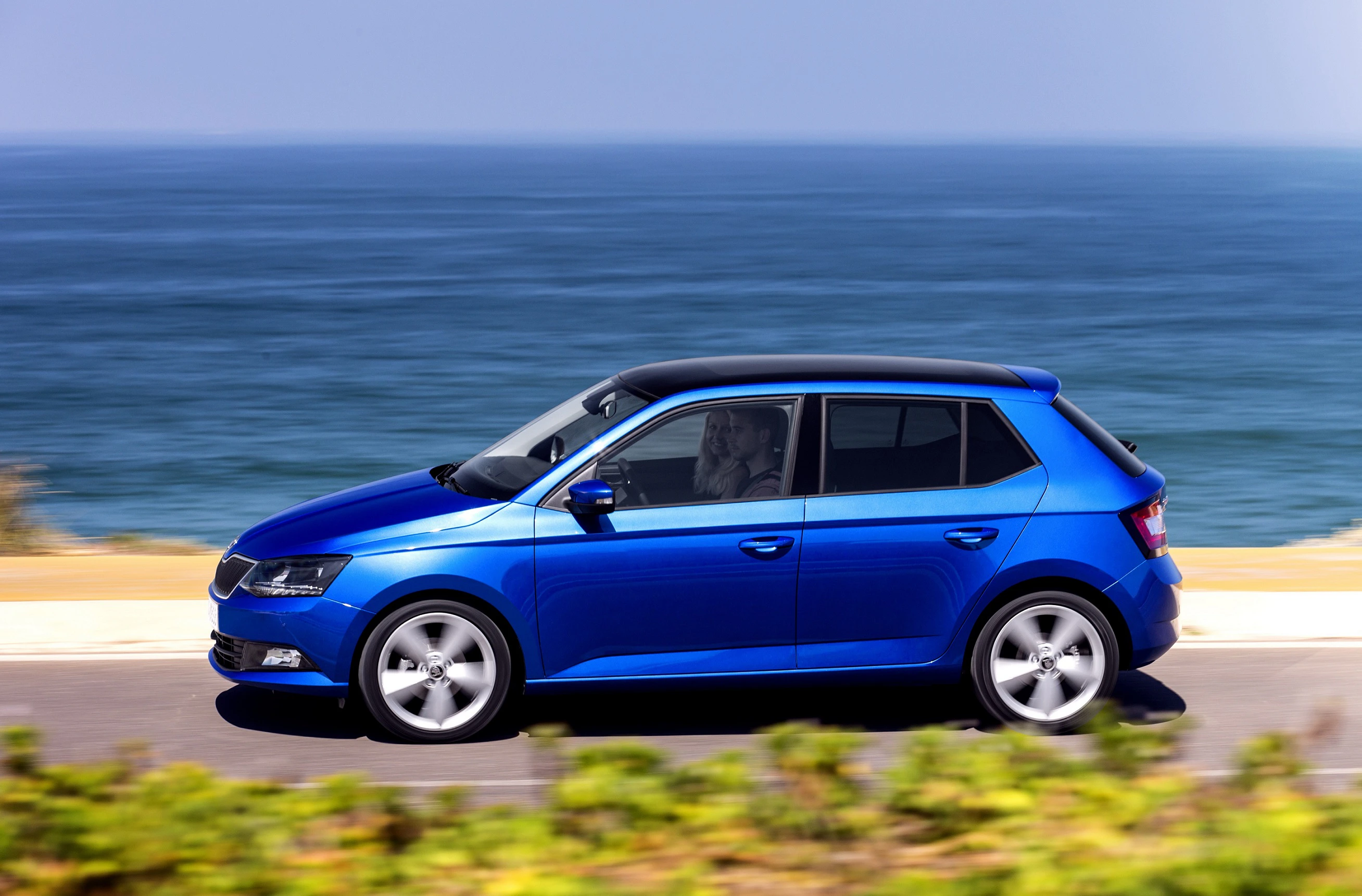 Blauer Kleinwagen fährt auf einer Küstenstraße entlang des Meeres; sonniger Tag mit klarem Himmel und grünem Bewuchs im Vordergrund.