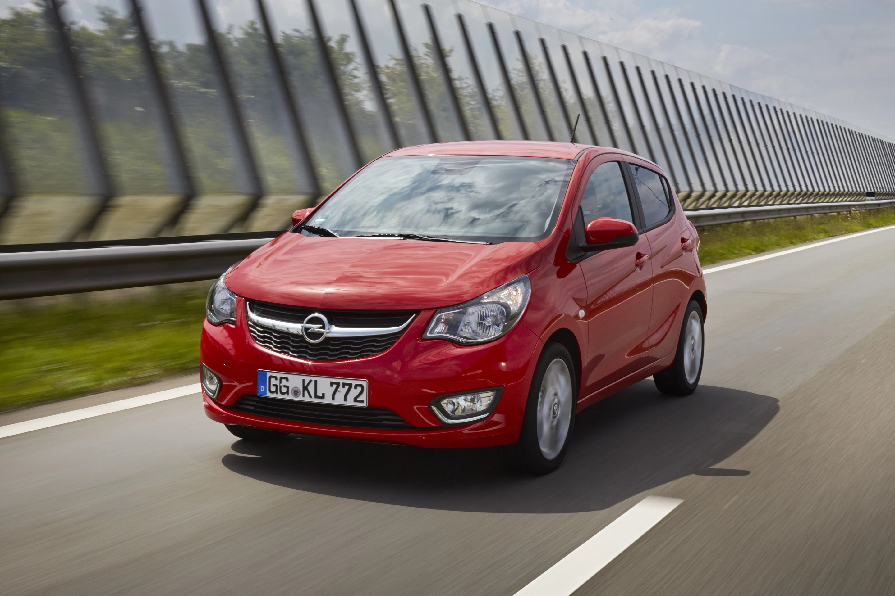 Roter Opel Kleinwagen fährt auf einer Autobahn, umgeben von Lärmschutzwänden, bewölkter Himmel im Hintergrund.