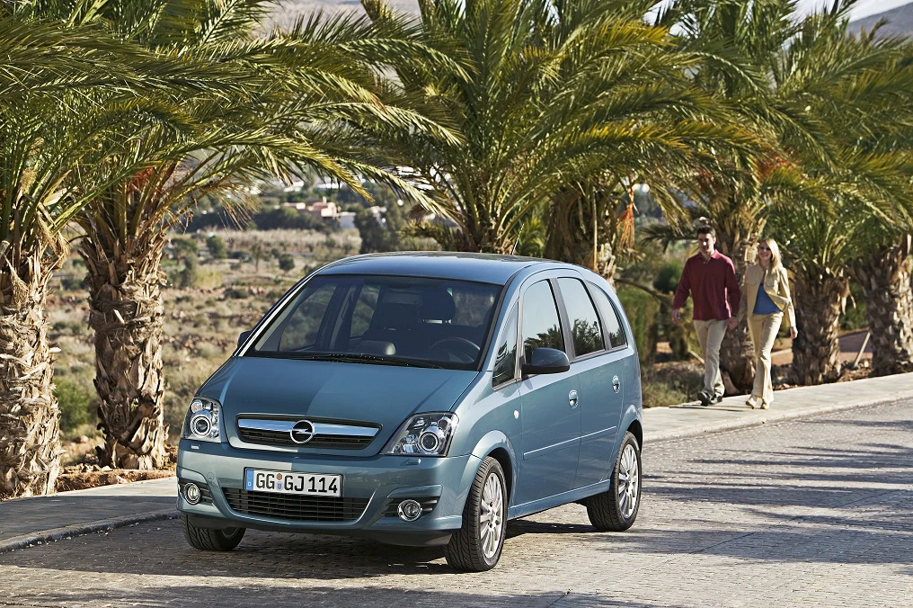 Blauer Opel-Van auf einer sonnigen Straße, gesäumt von Palmen; im Hintergrund geht ein Paar spazieren.