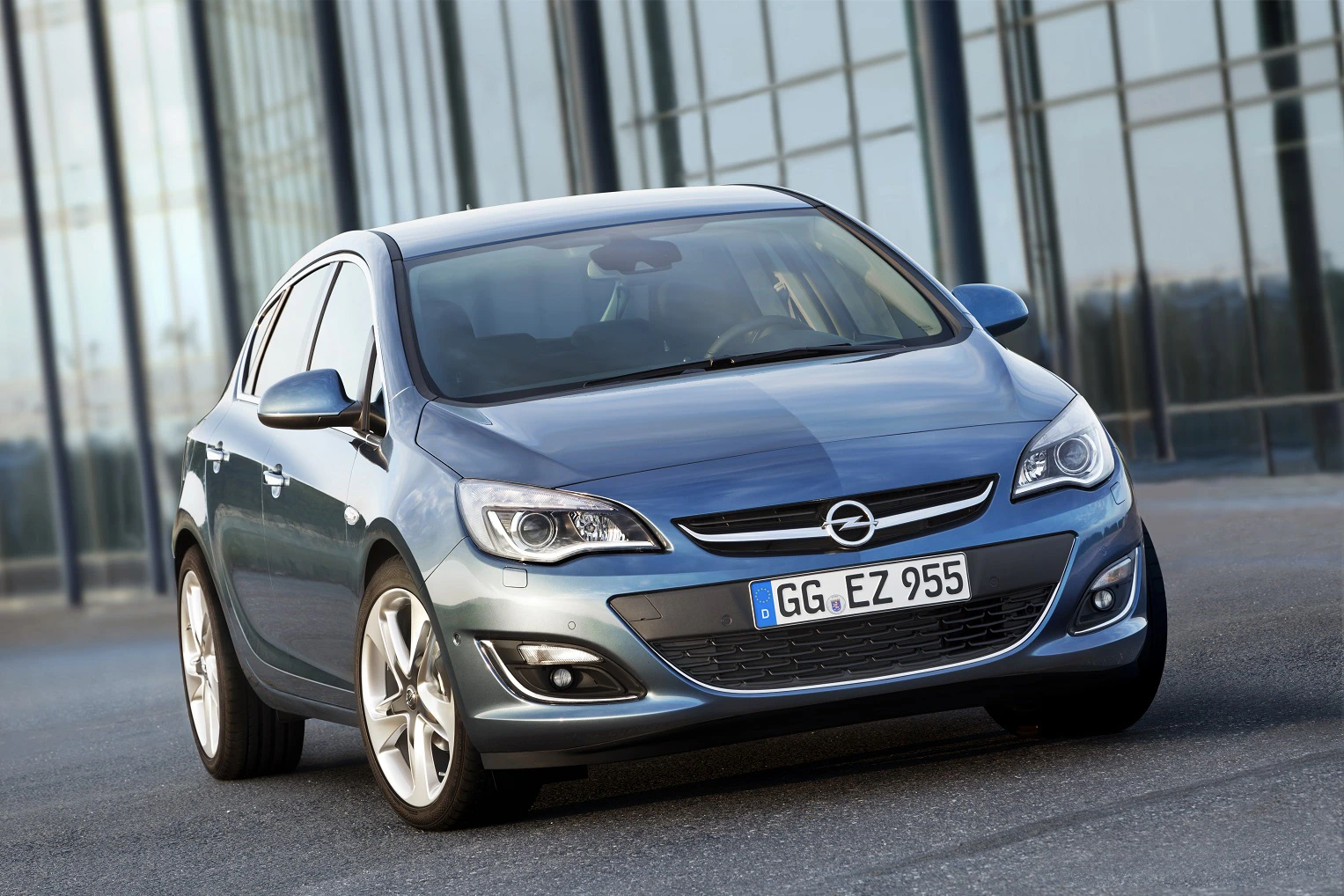 Blauer Opel Astra, geparkt vor modernen Glasgebäuden. Lichter reflektieren auf der Karosserie, Auto zeigt Frontansicht.