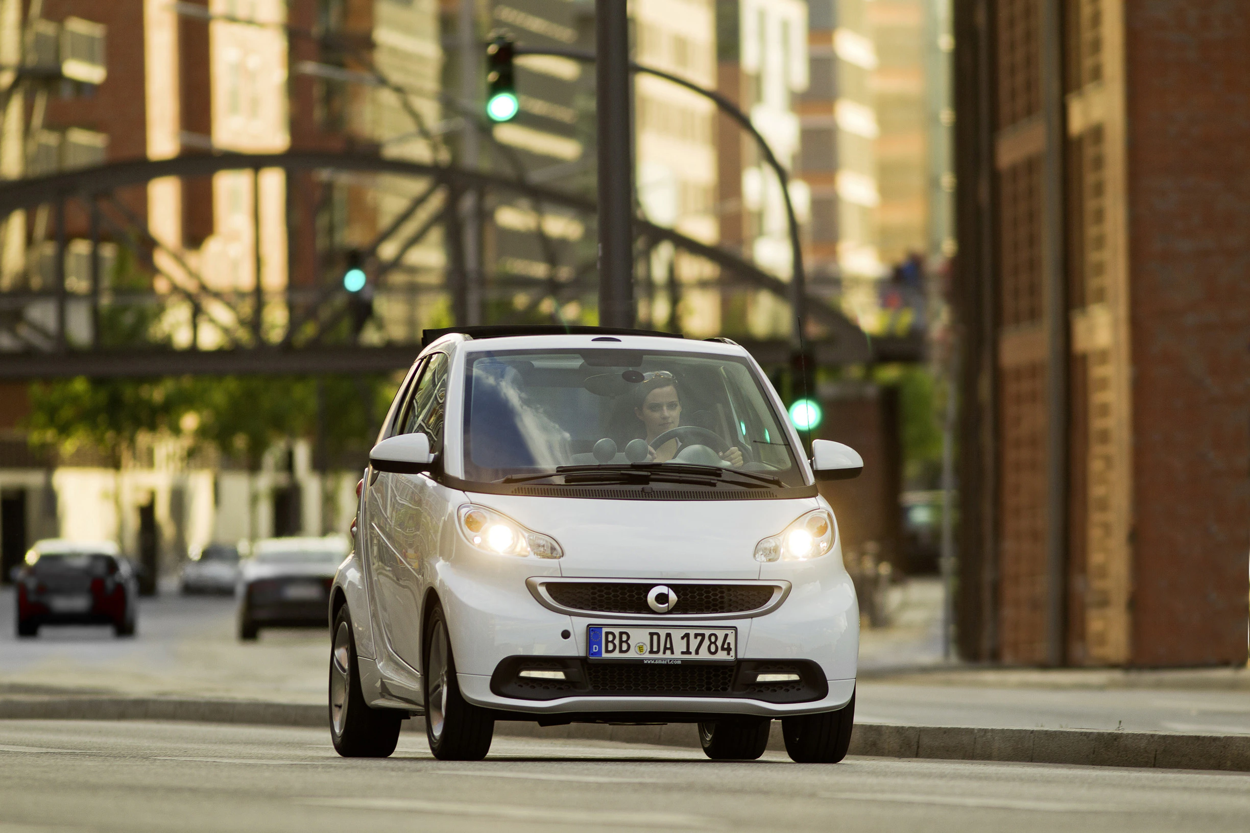 Weißer Smart fährt auf einer Stadtstraße, in Bewegung. Im Hintergrund sind Gebäude und eine grüne Ampel sichtbar.