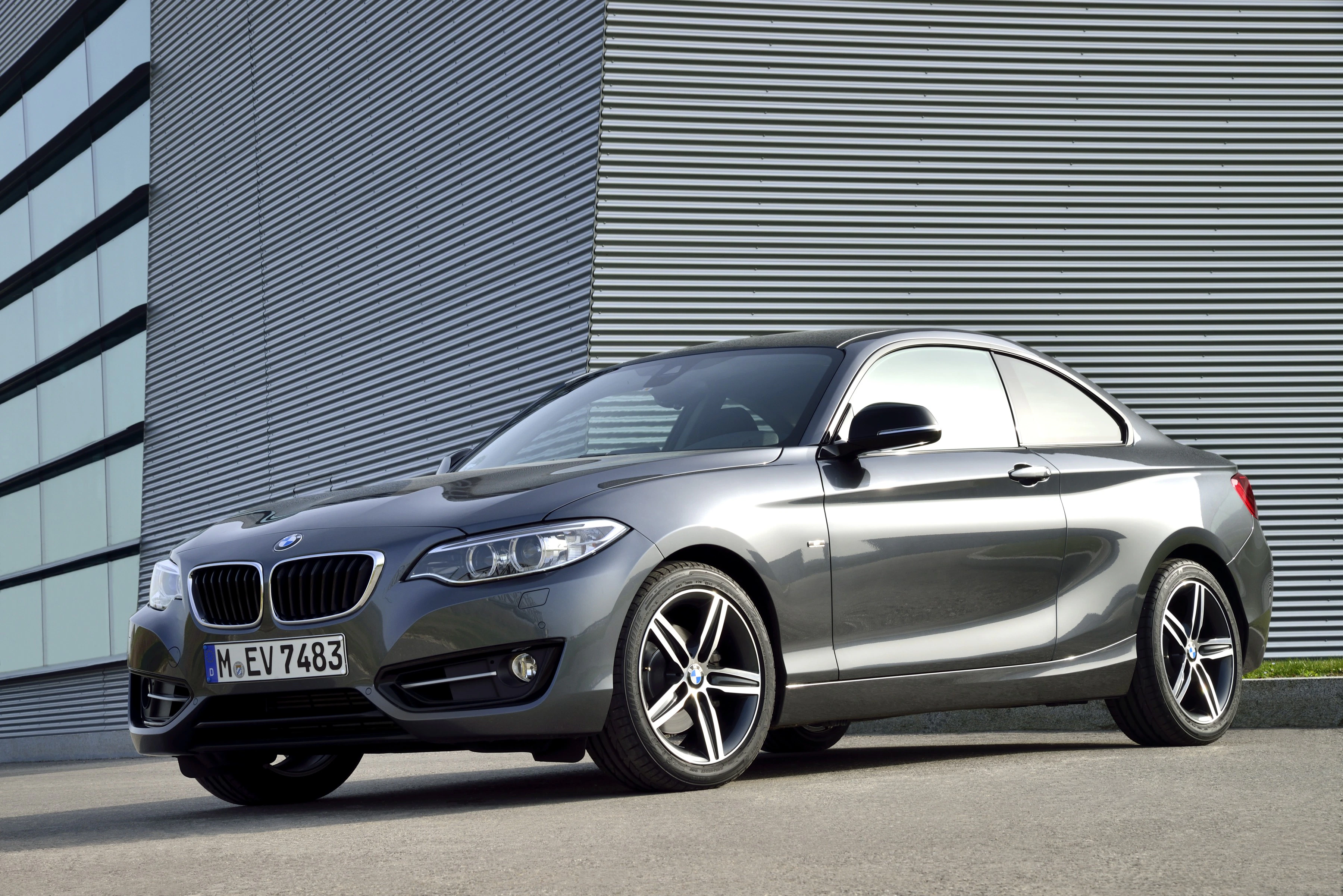 Graues BMW Coupé steht vor einem modernen Gebäude mit horizontalen Linien und großen Fenstern. Fahrzeug zeigt dynamisches Design.