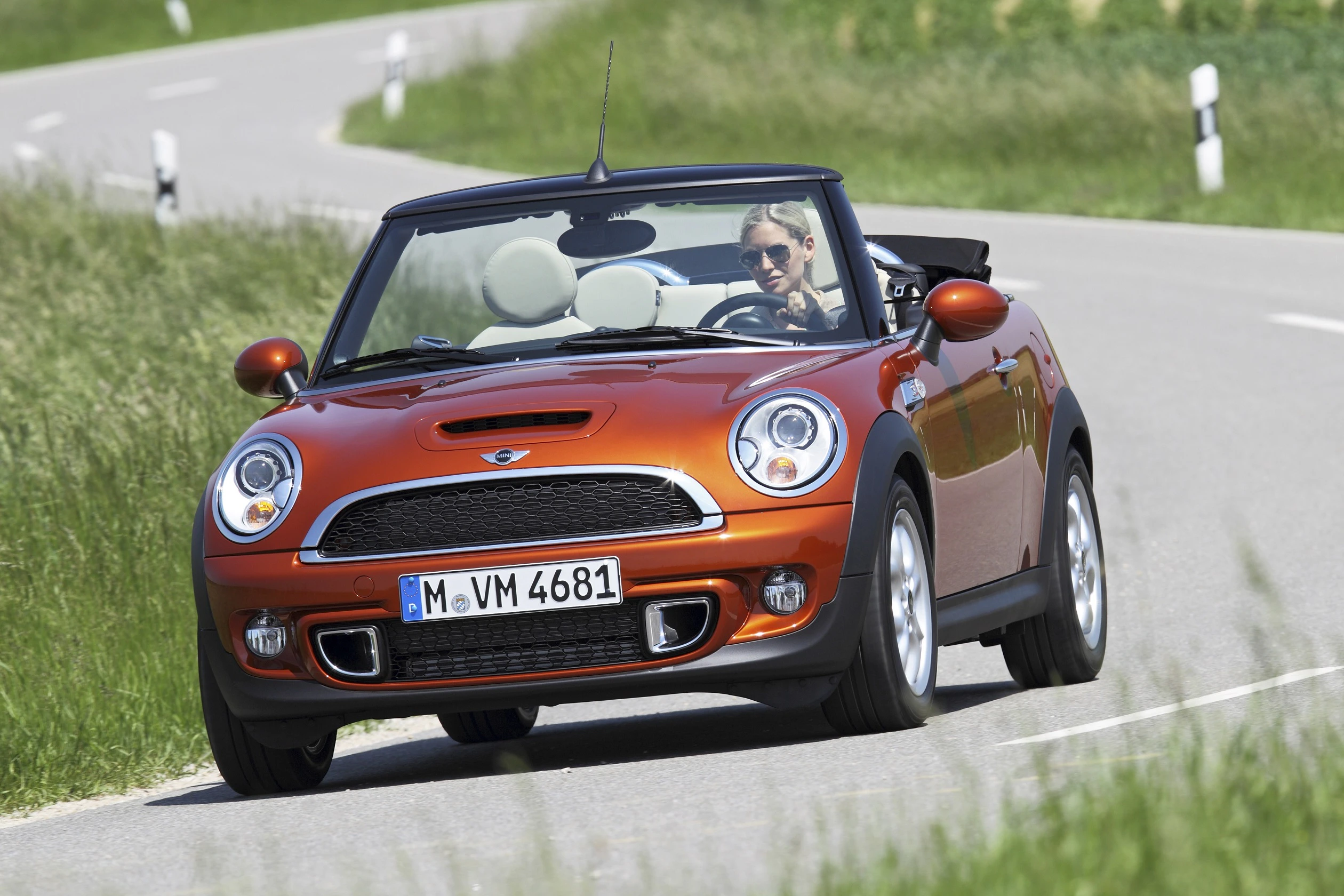 Oranges Cabrio fährt auf einer kurvigen Landstraße bei sonnigem Wetter. Fahrer trägt Sonnenbrille. Umgeben von grüner Landschaft.