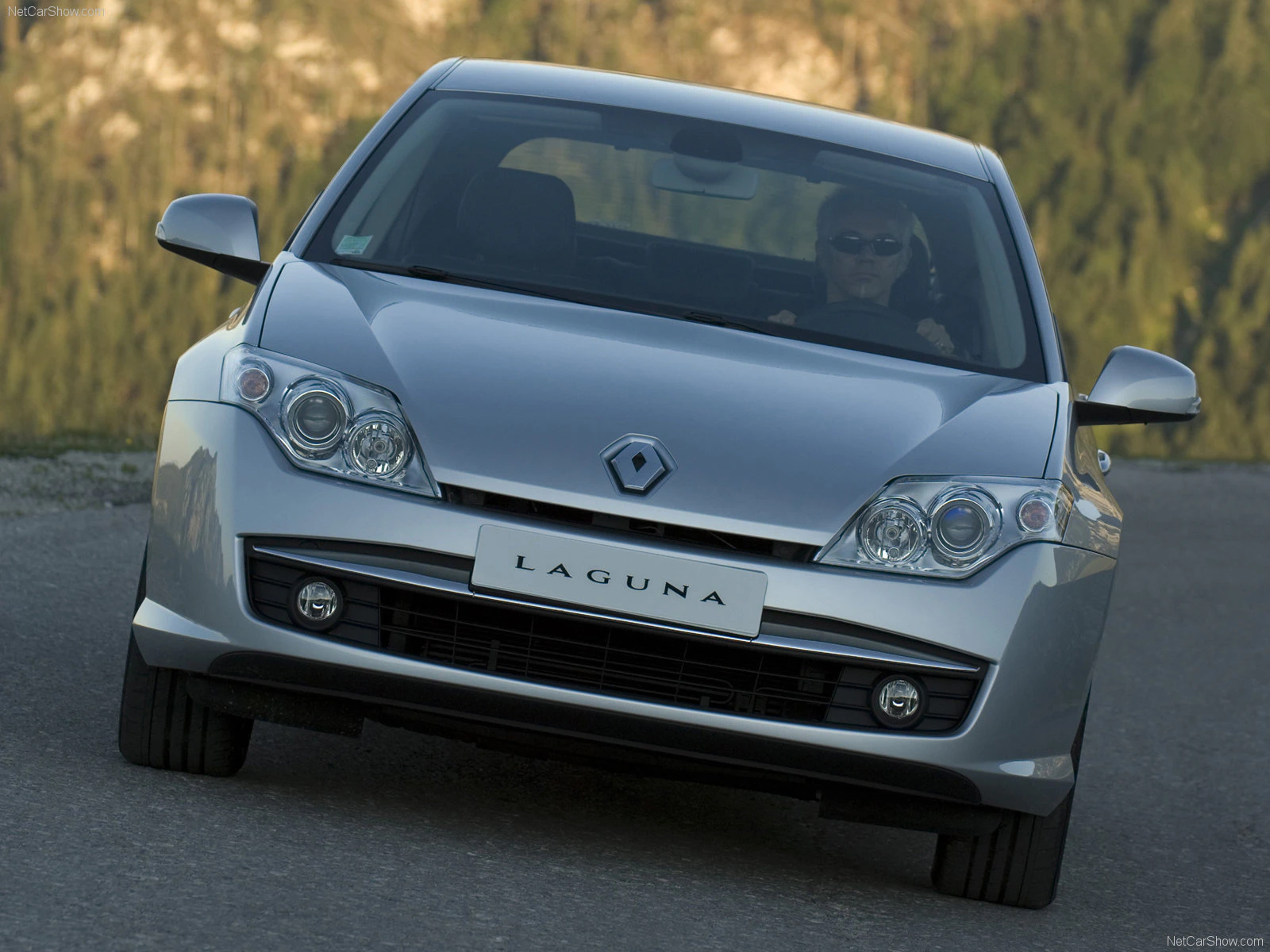 Silberner Renault Laguna fährt auf einer Straße, mit Bäumen im Hintergrund. Fahrer mit Sonnenbrille am Steuer.