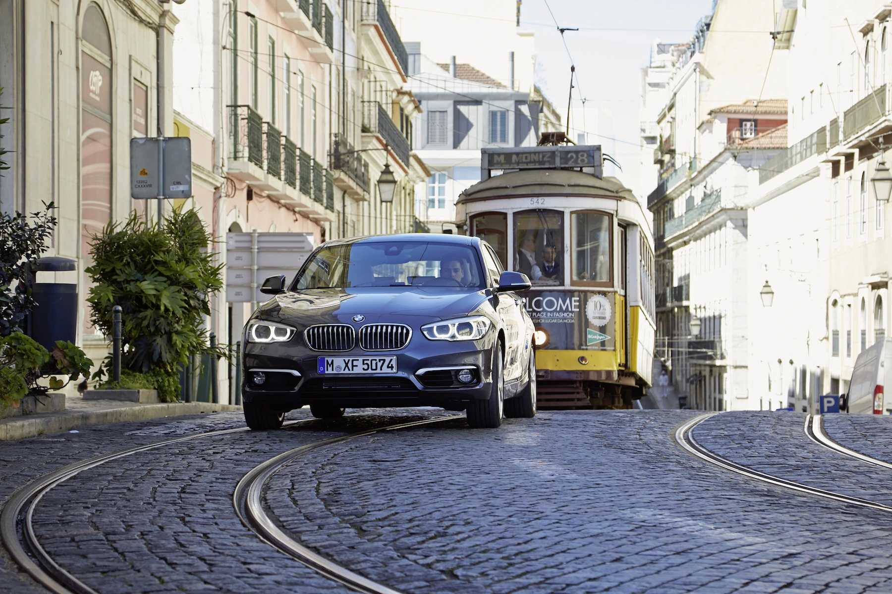 Ein schwarzer BMW fährt auf kopfsteingepflasterter Straße, während sich eine gelbe Straßenbahn nähert. Historische Gebäude im Hintergrund.