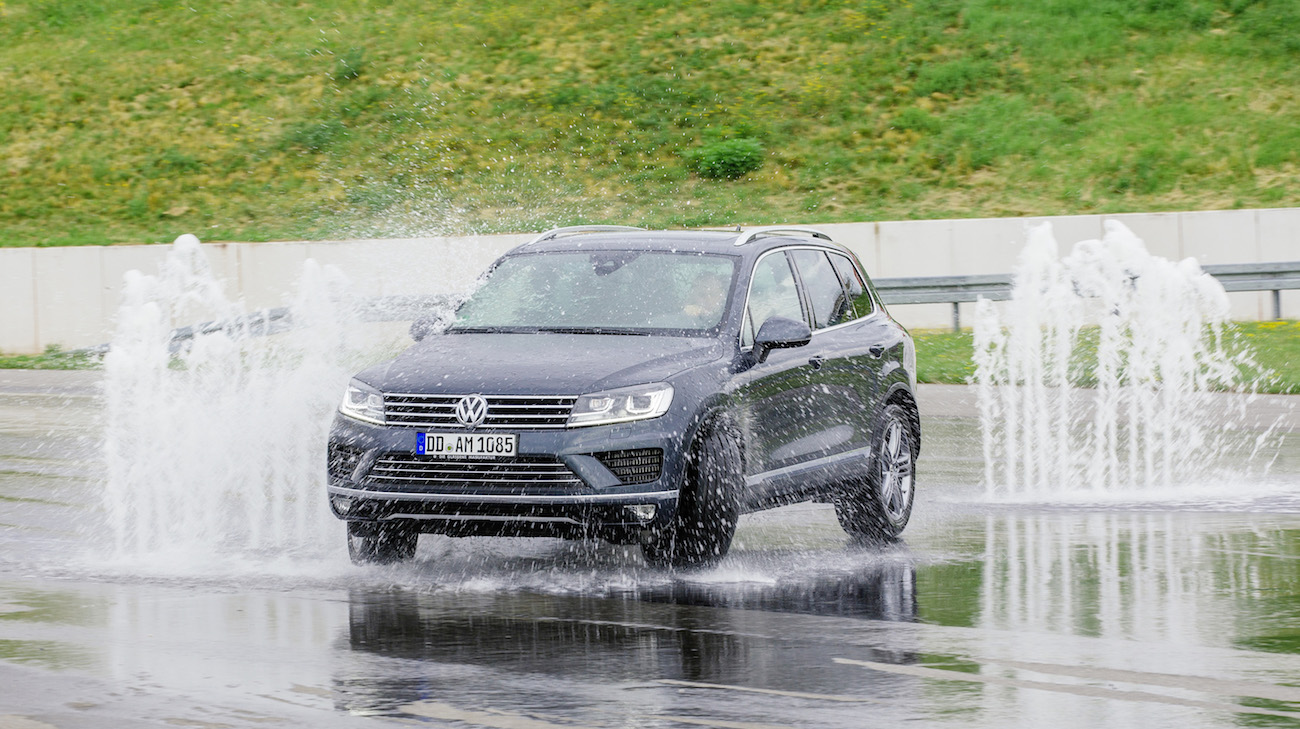 Ein grauer SUV fährt durch eine Wasserfontäne auf einer nassen Teststrecke.