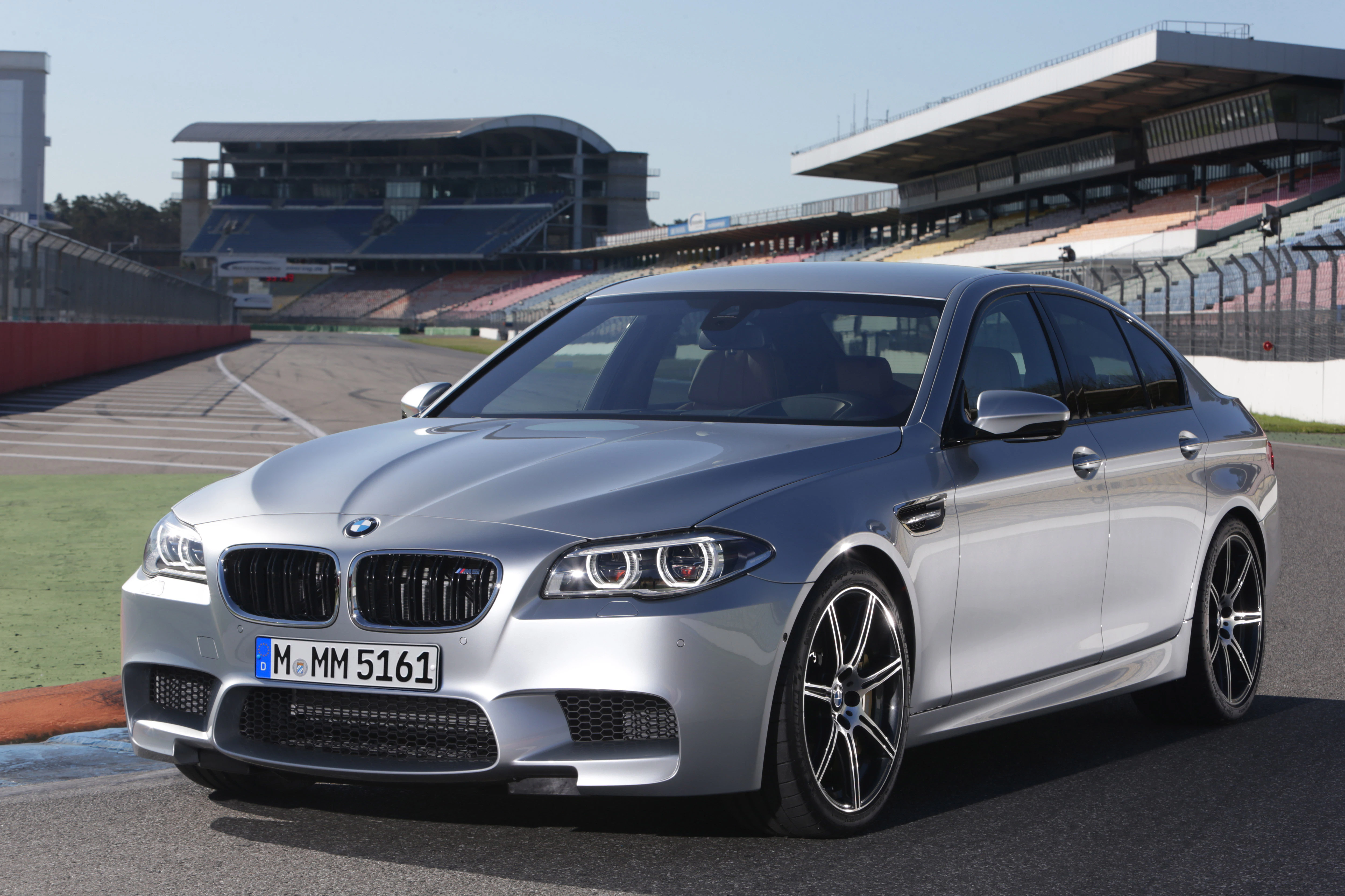 Silberner BMW auf einer Rennstrecke bei Tageslicht, Gebäude im Hintergrund. Sportliches Design mit auffälligen Felgen.