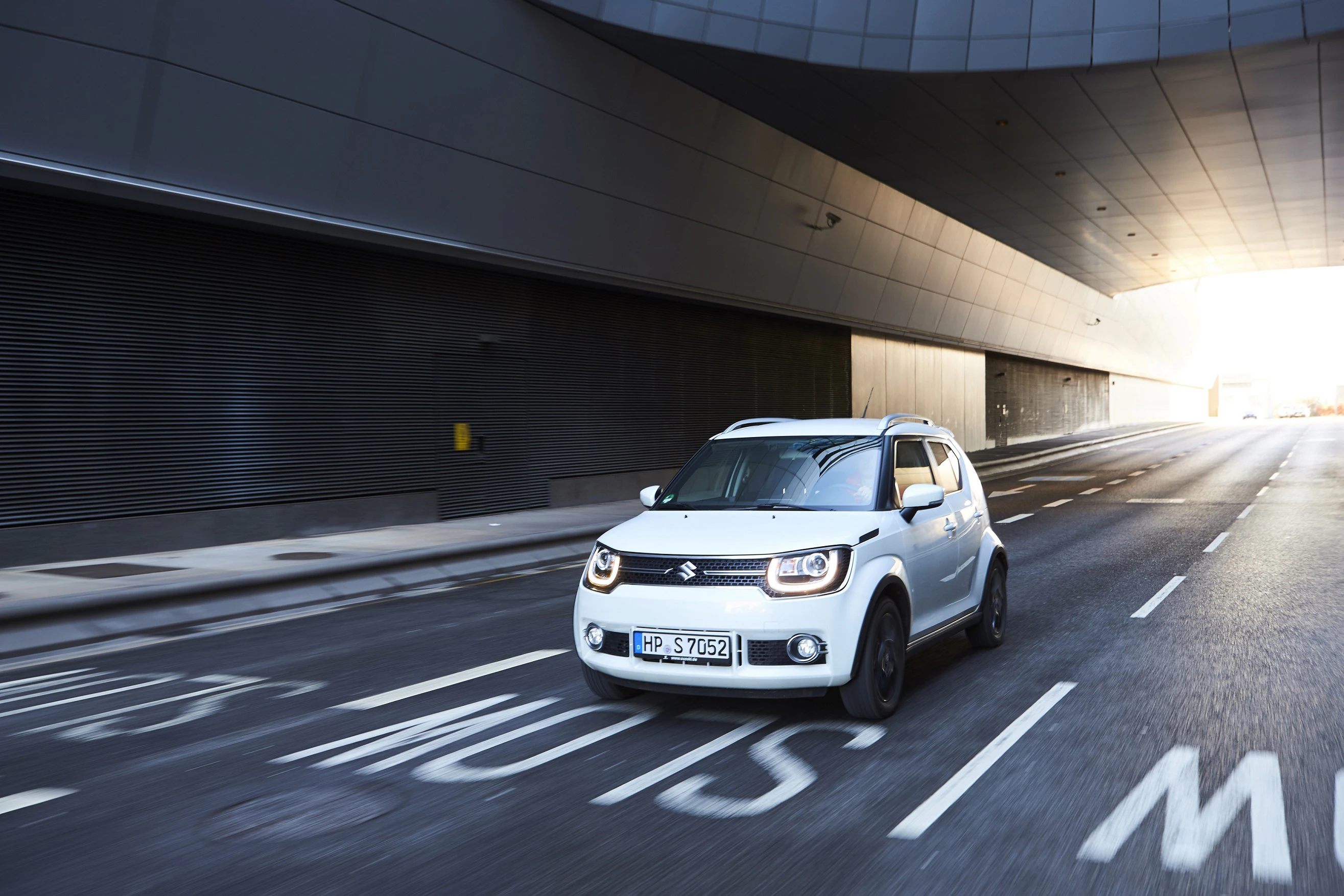 Ein weißer Suzuki fährt auf einer leeren, modernen Straße durch einen Tunnel. Sonnenschein erhellt den Hintergrund.