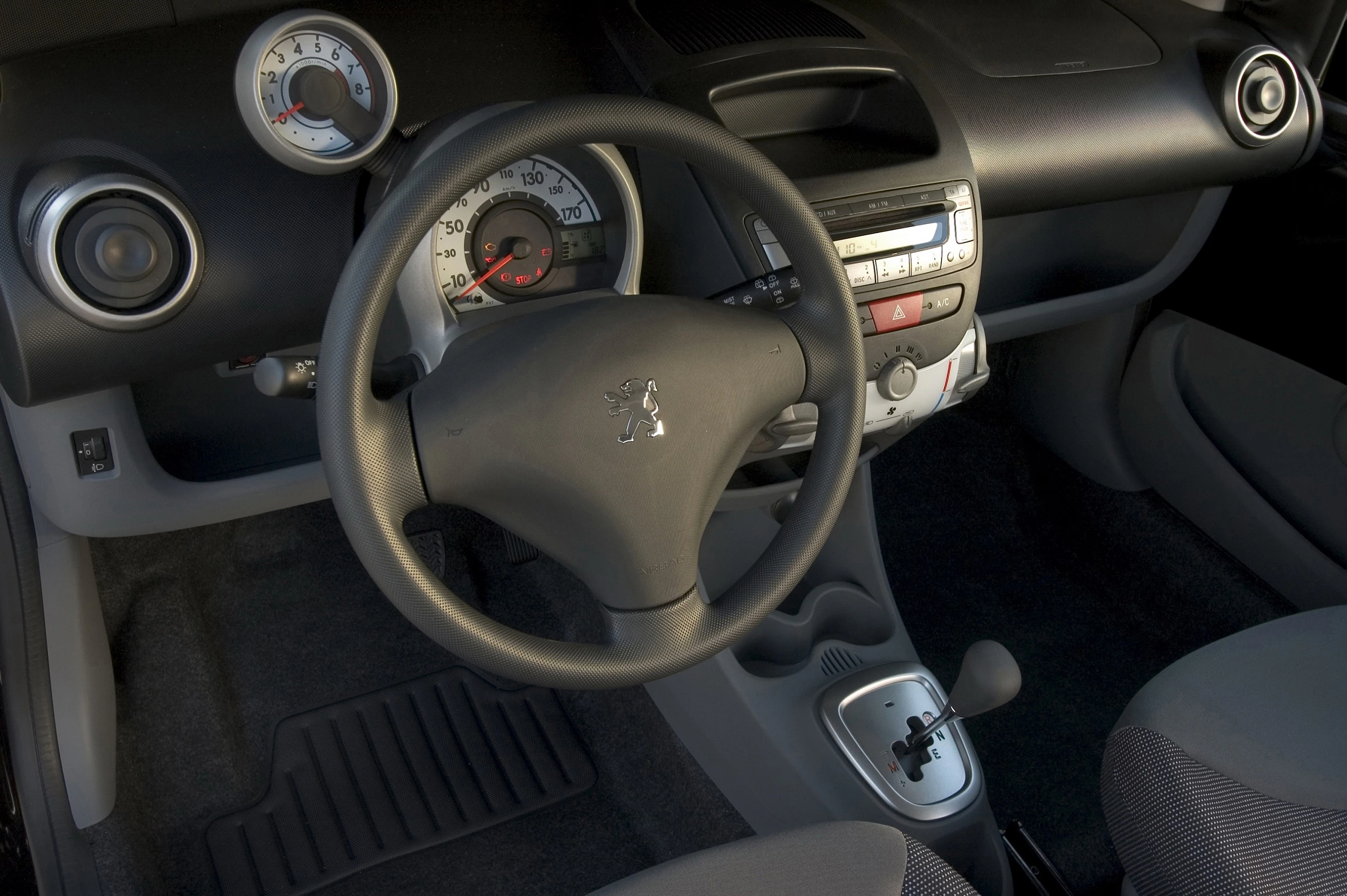 Innenraum eines Autos mit Peugeot-Logo auf dem Lenkrad, Tacho und Radiosystem im Cockpit, Automatikschaltung und grauen Sitzen.