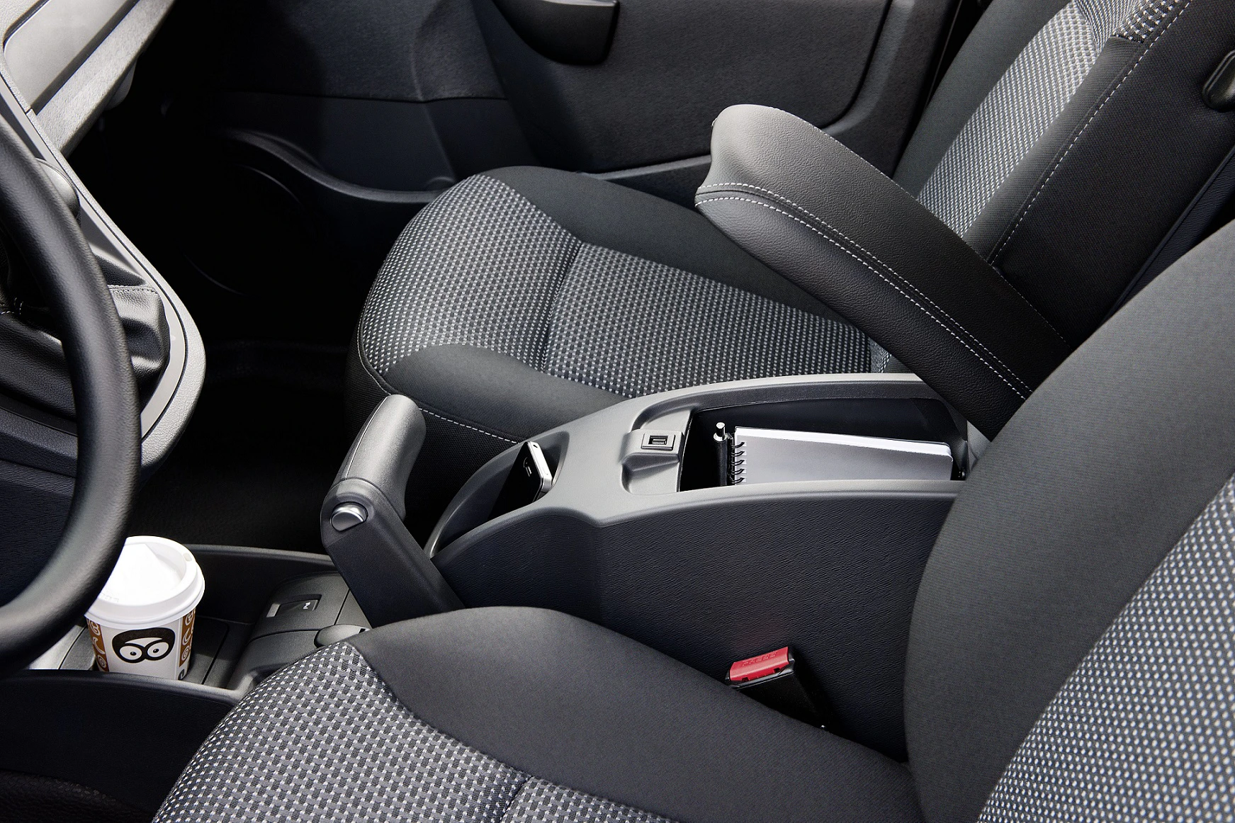 Innenraum eines Autos mit zwei grauen, gepolsterten Sitzen, Mittelkonsole mit Stauraumfach, Getränkehalter mit Kaffeebecher und Sicherheitsgurten.